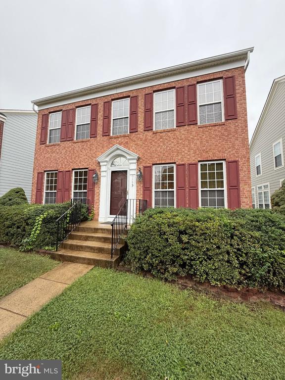 Two-story red brick house with burgundy shutters, front steps, and trimmed shrubs.