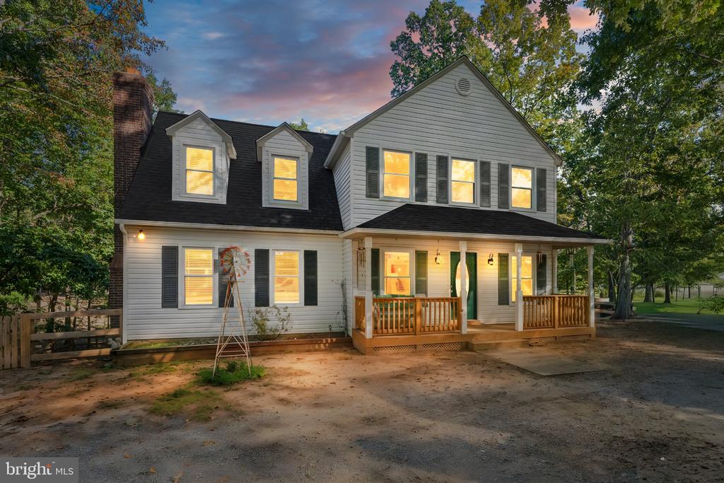 Two-story white house with lit windows, dormers, and a covered wooden porch at dusk.