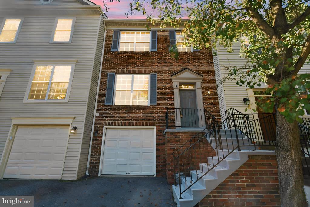 Brick townhouse with white siding, garages, blue shutters, and a raised entrance framed by a leafy tree.