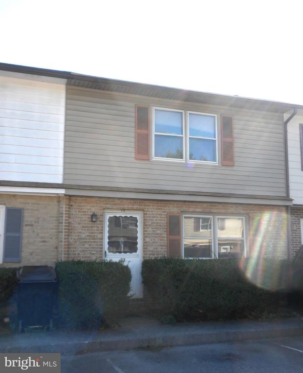 Two-story attached home with tan siding, brick lower level, shuttered windows, and front hedges.
