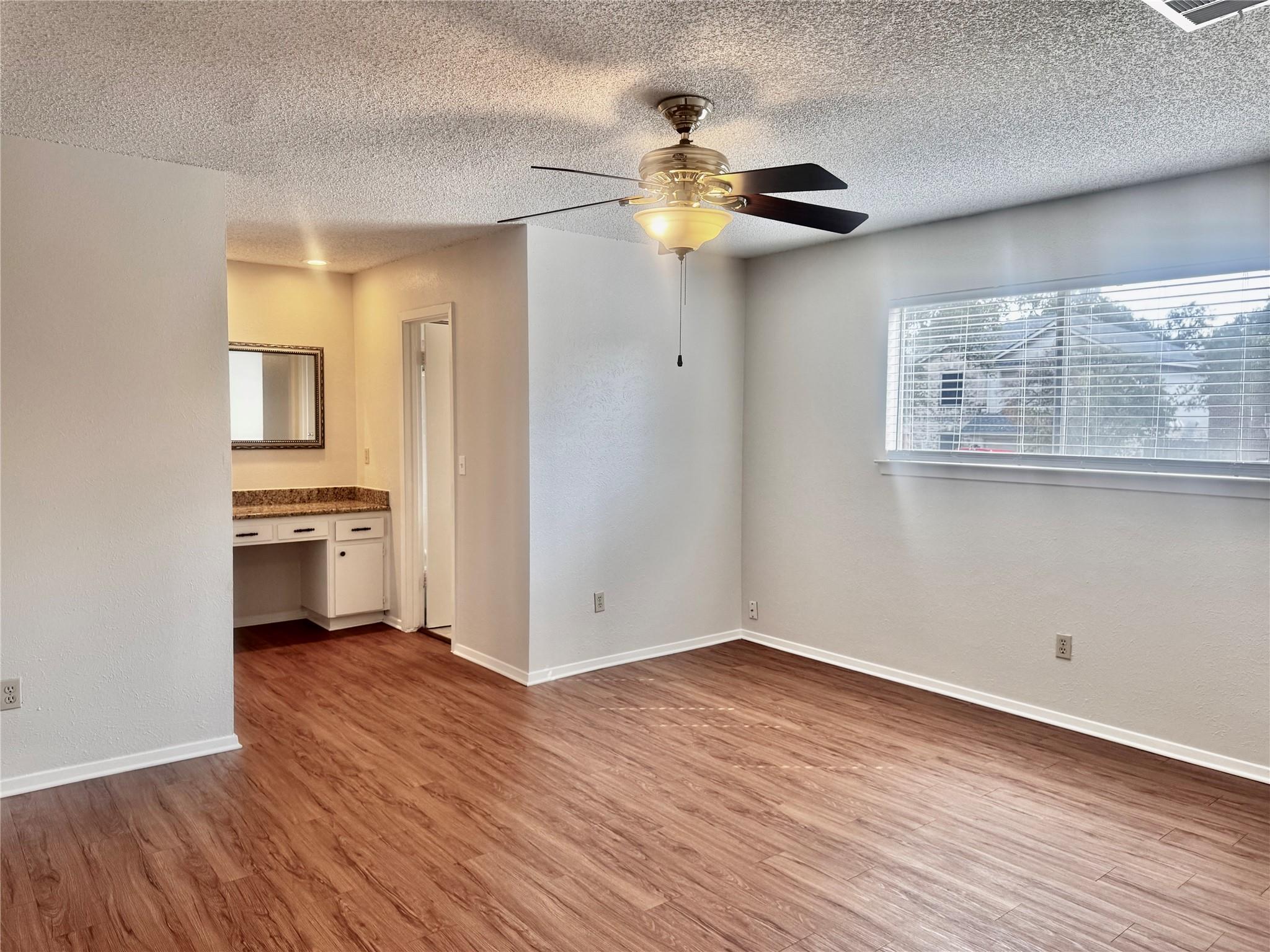 Vacant bedroom with wood-look flooring, ceiling fan, window blinds, and an adjoining vanity area.