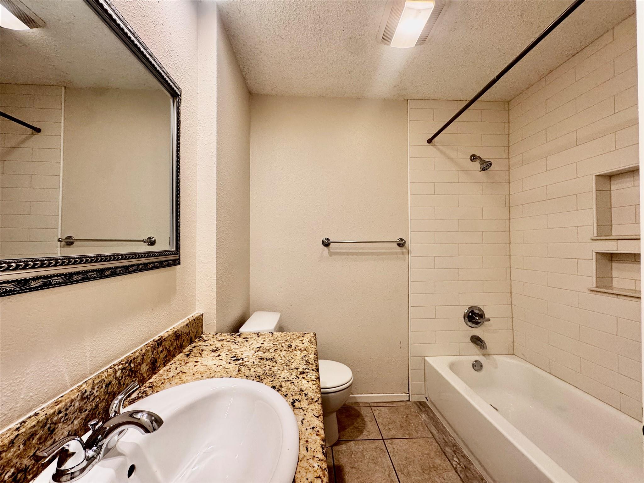 Bathroom with a white tiled tub and shower beside a granite vanity and large framed mirror.