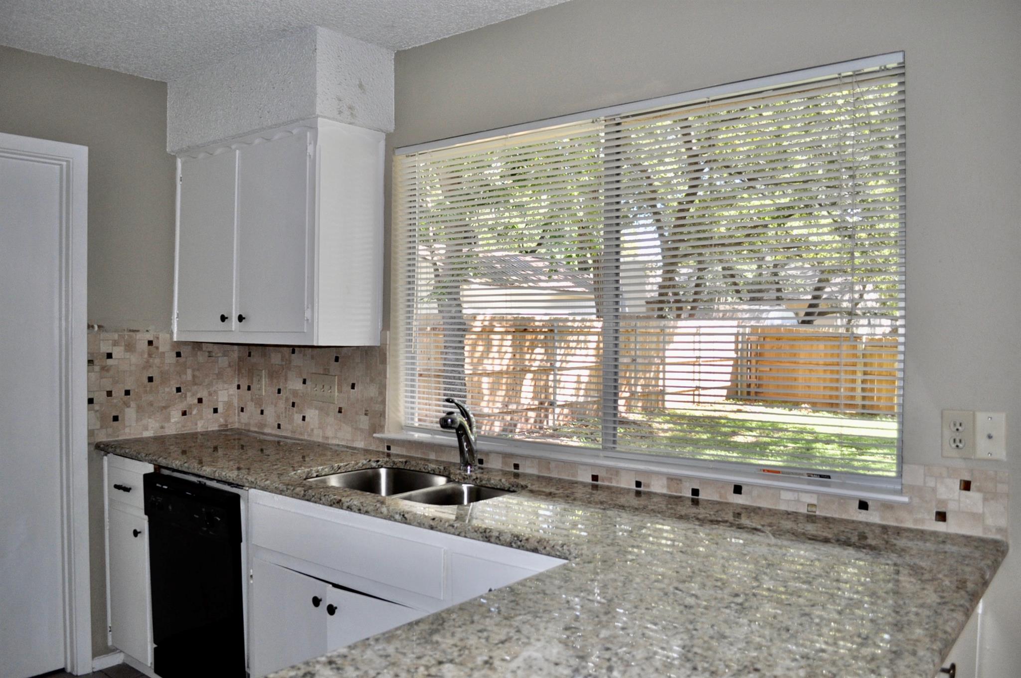 Kitchen with granite counters, white cabinets, a double sink, and a large window with blinds.