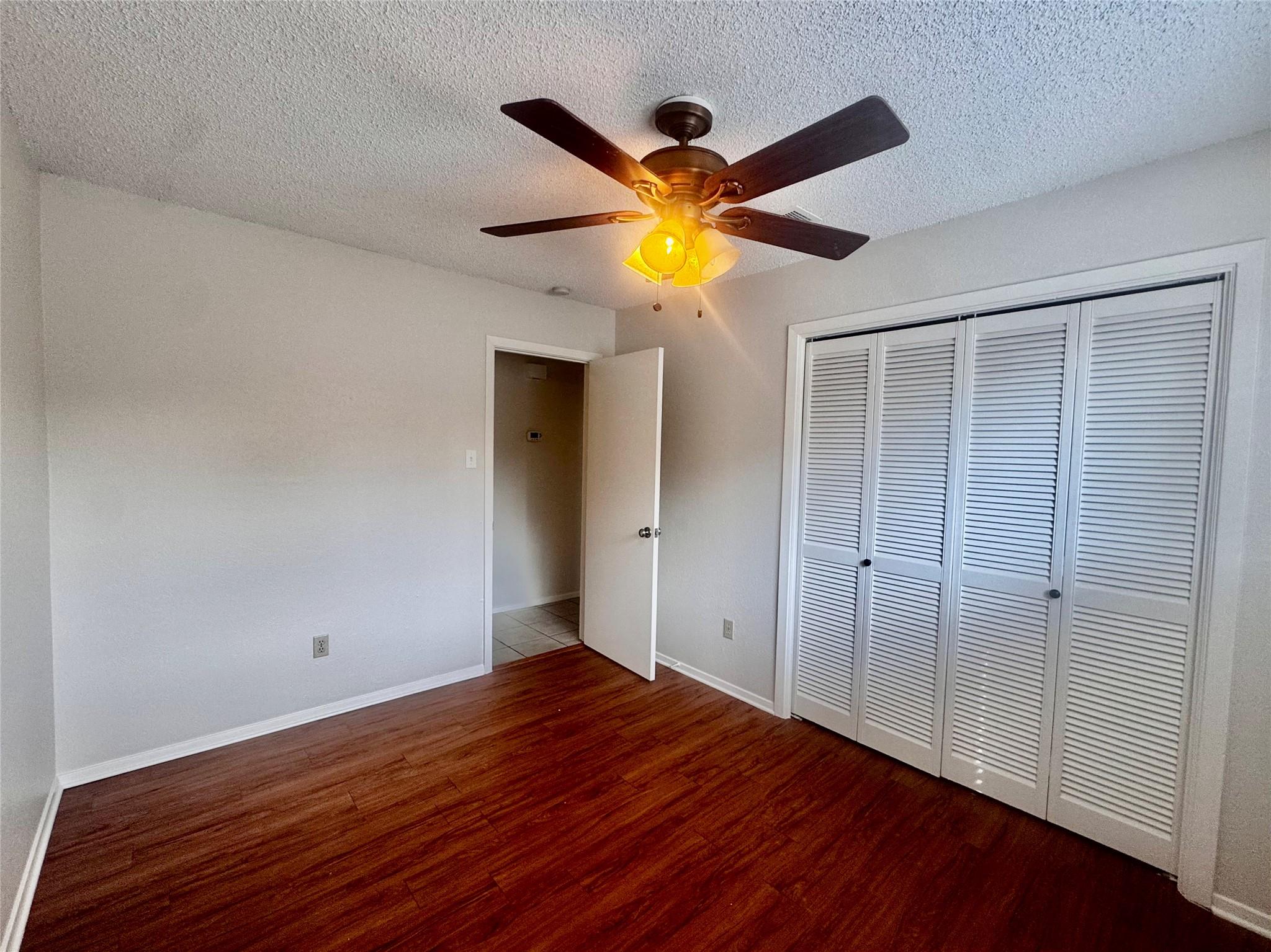 Empty bedroom with wood flooring, louvered closet doors, and a lit ceiling fan.