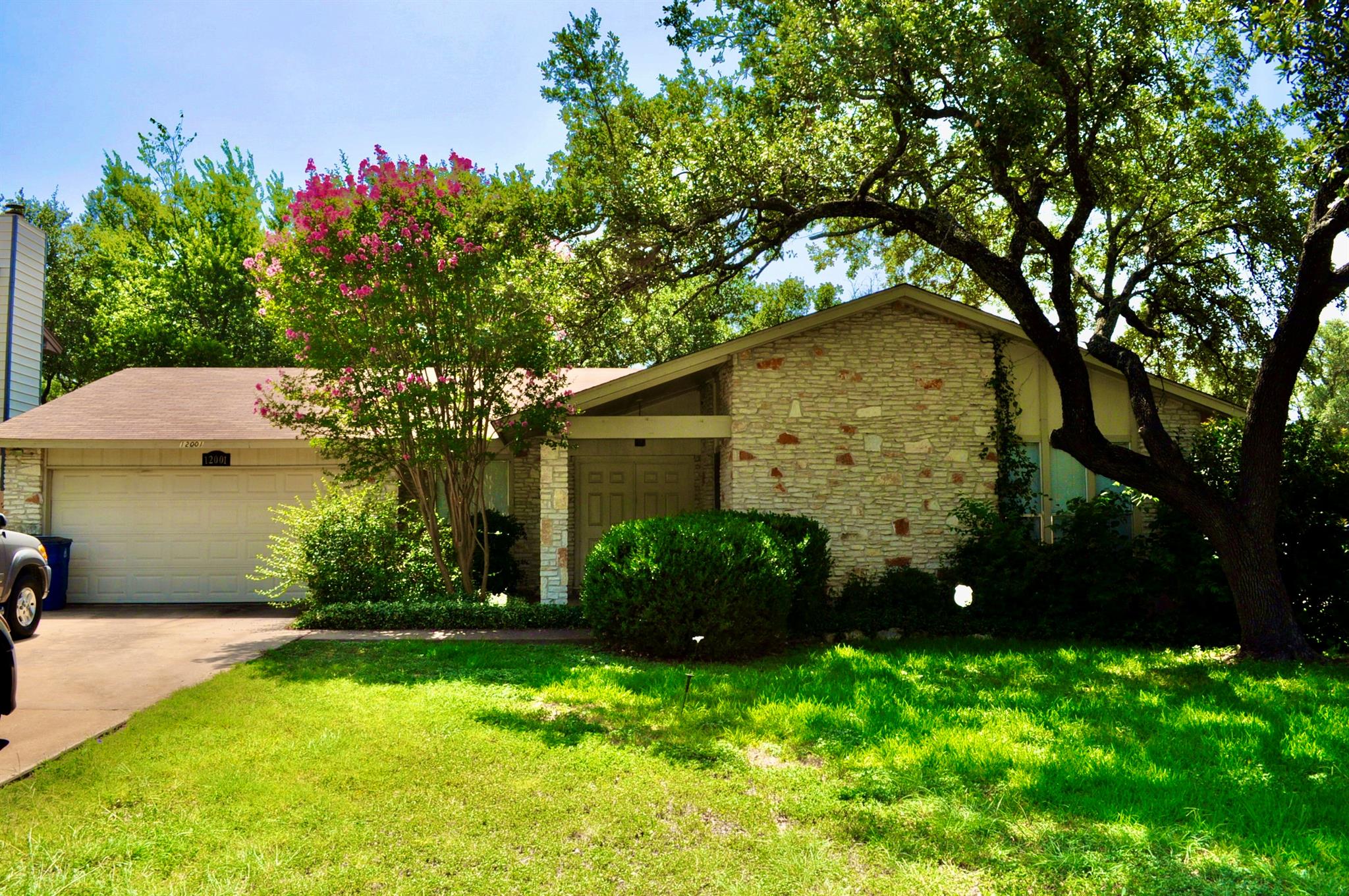 Single-story stone-front home with an attached garage, shaded lawn, and pink-flowering tree.