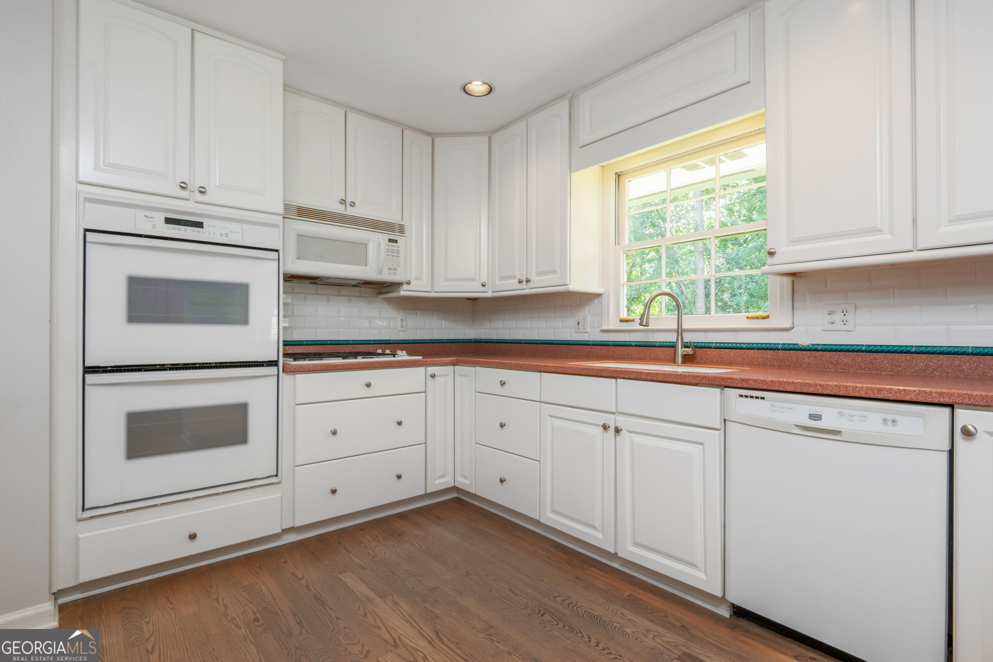 Kitchen with white cabinetry, double ovens, reddish counters, and a window above the sink.