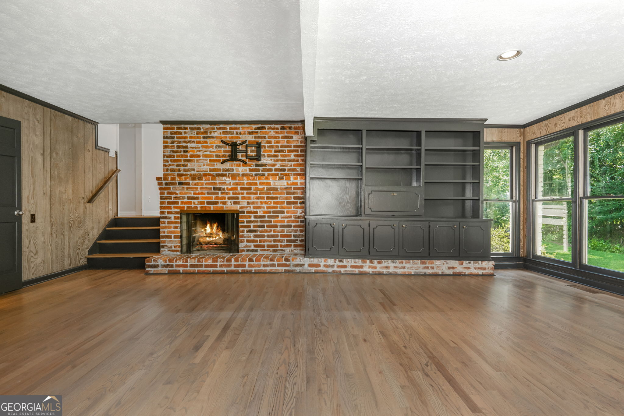 Vacant living room with a brick fireplace, dark built-ins, hardwood floors, and large windows.