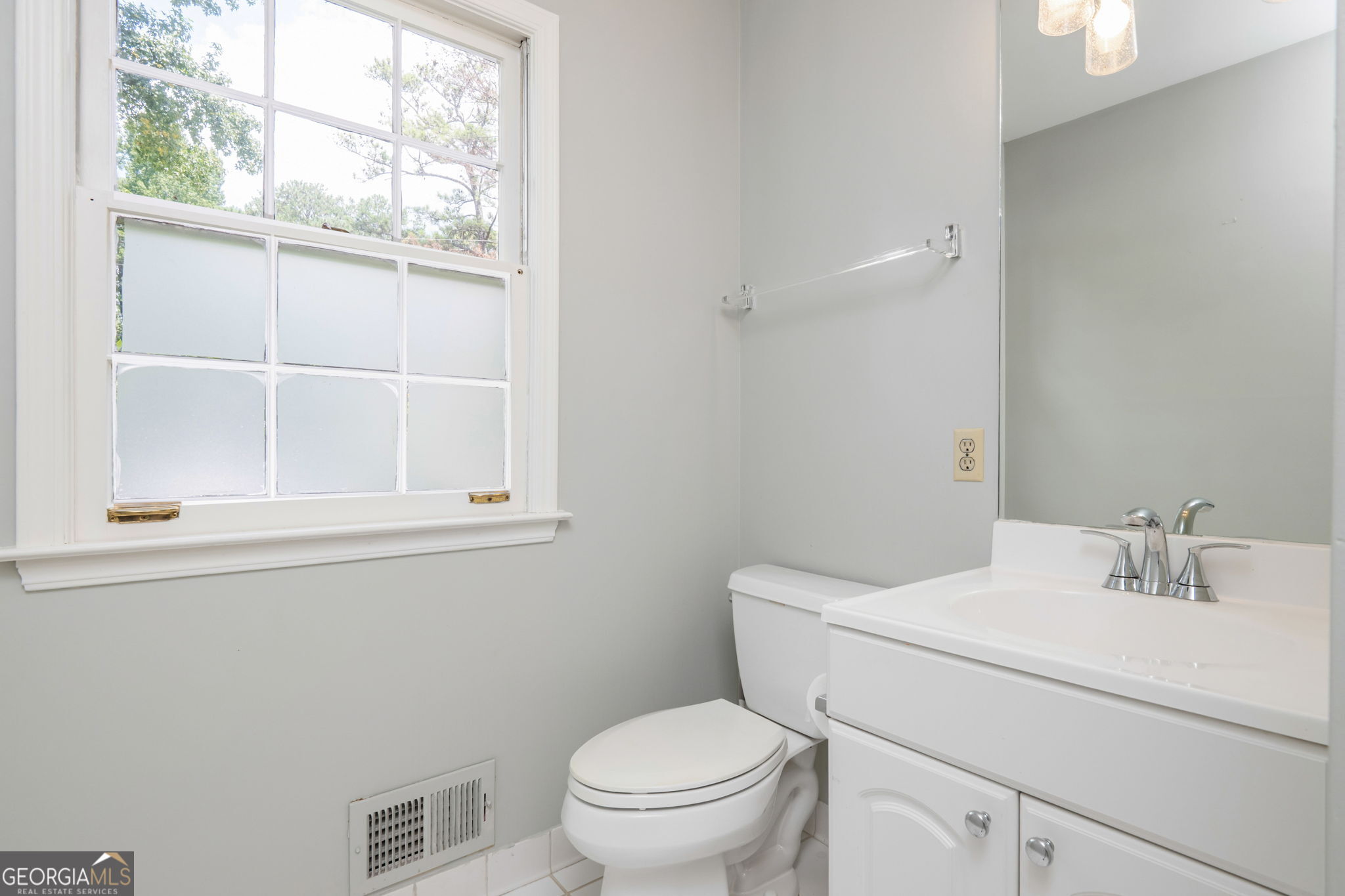 Bathroom with a white vanity and toilet beside a large window with frosted panes.