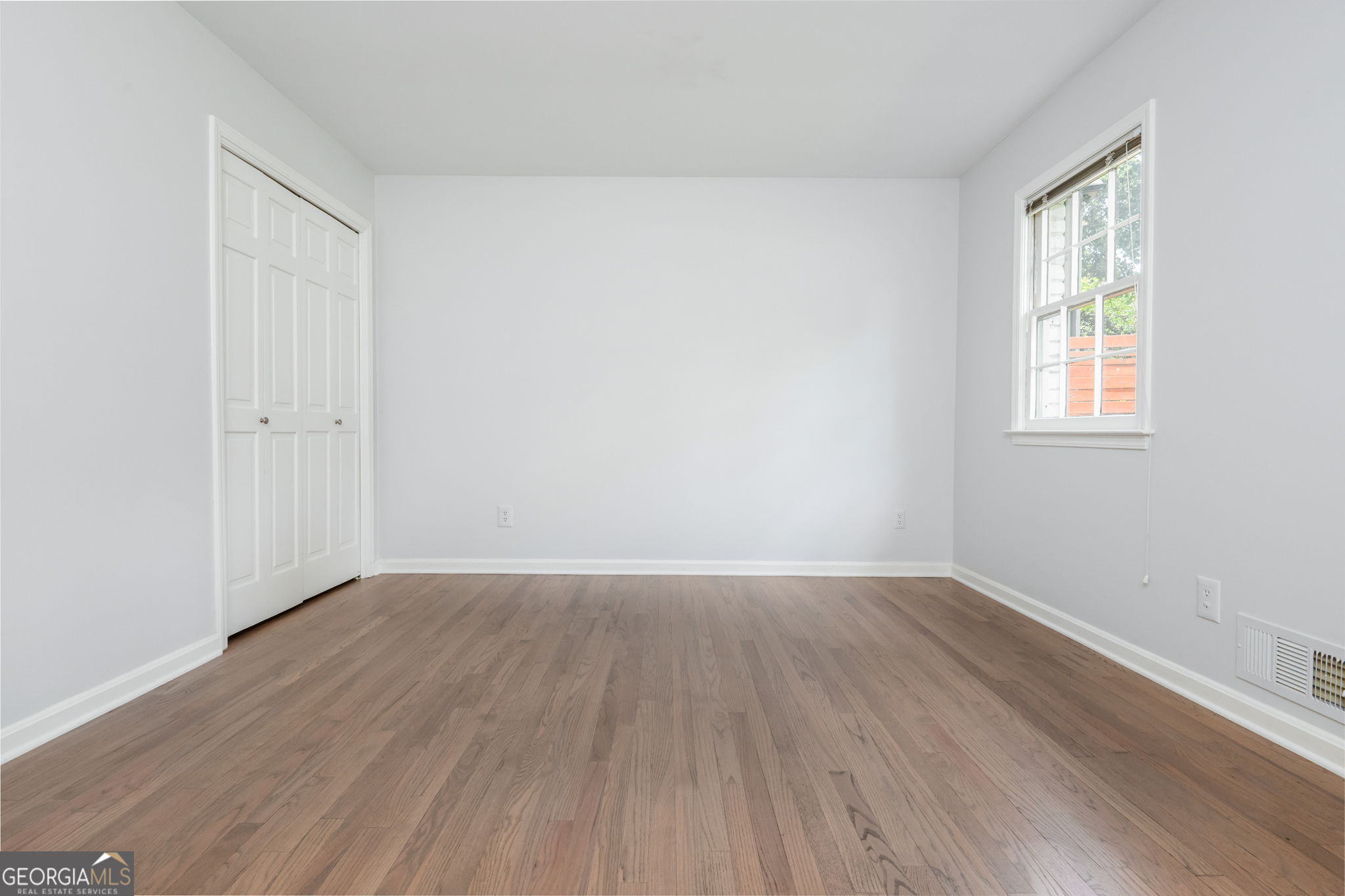 Empty bedroom with hardwood floors, a double-door closet, and a sunlit window.