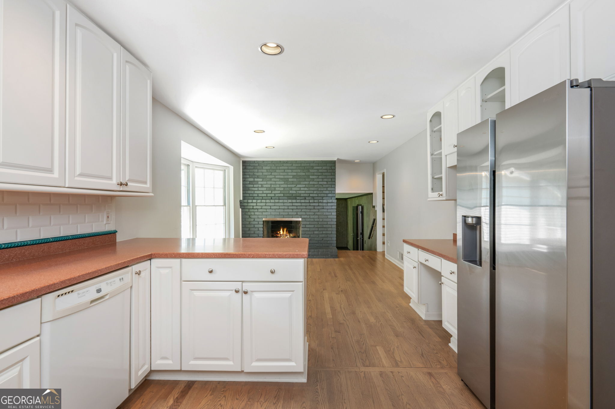 Kitchen with white cabinets and reddish counters opening toward a green brick fireplace.