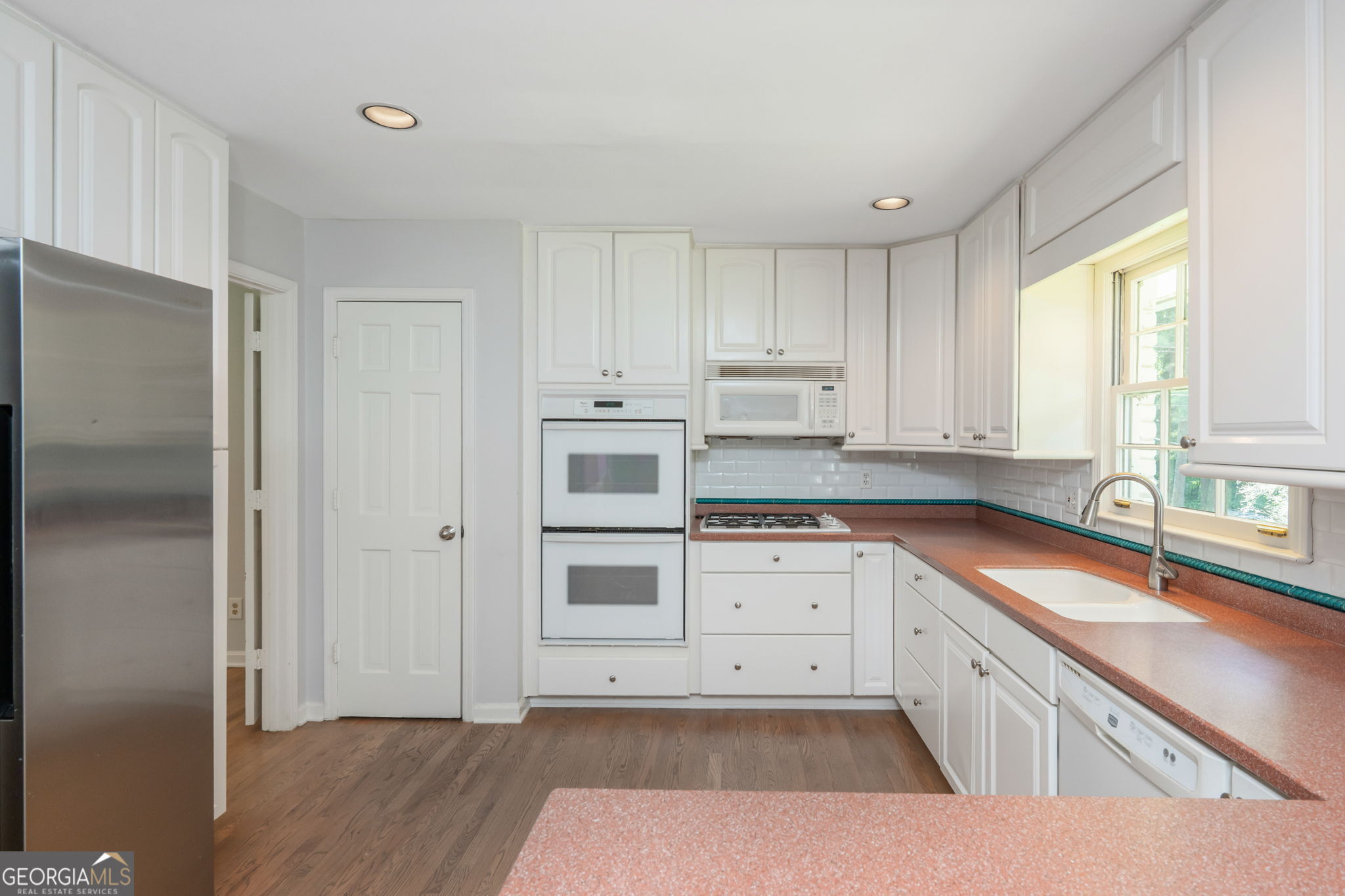 Kitchen with white cabinets, double wall oven, reddish countertops, and a sink beneath a bright window.