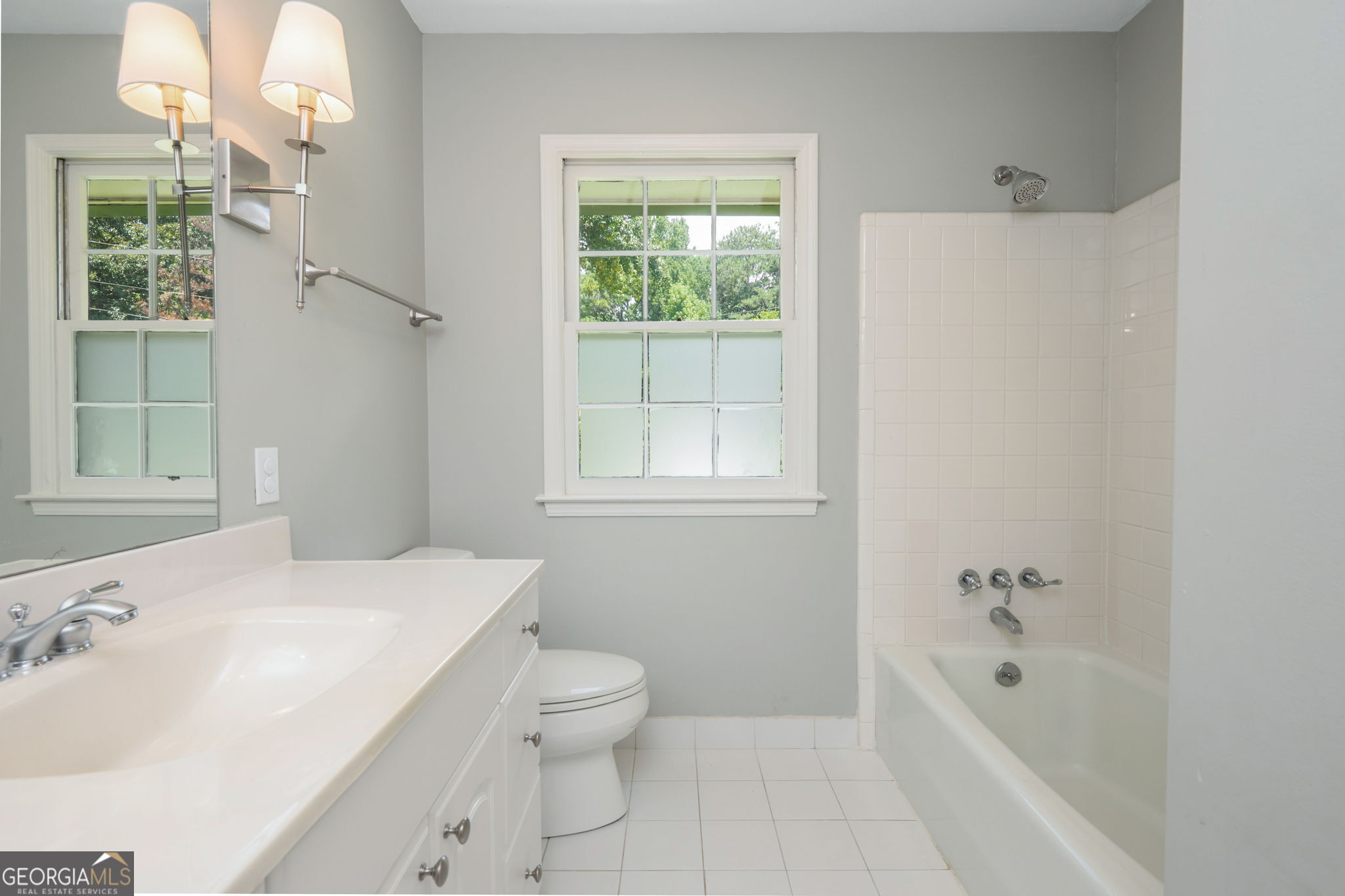 Bathroom with white vanity, tiled tub and shower, and two daylight-filled windows.