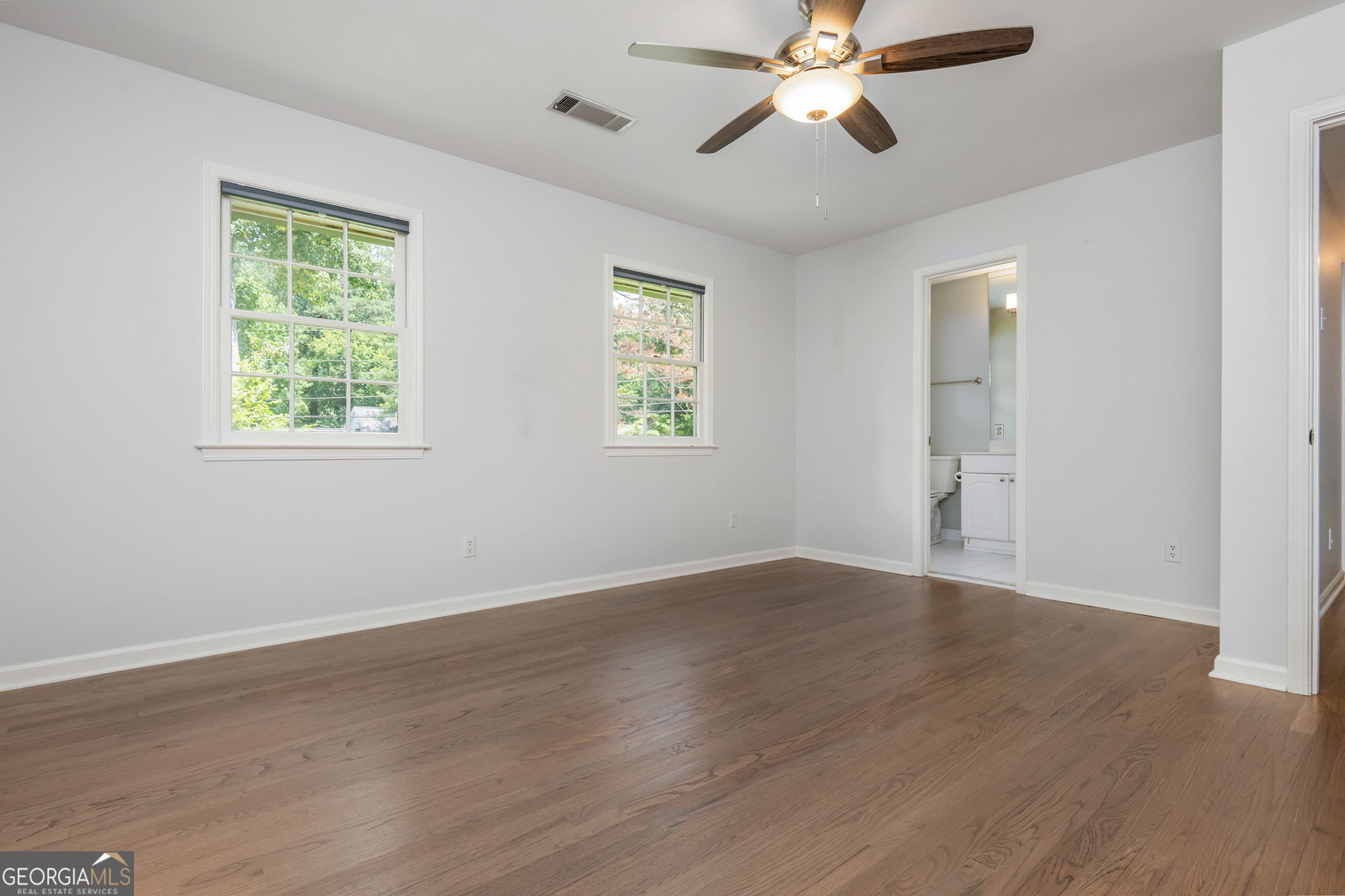 Empty bedroom with hardwood floors, two windows, and a ceiling fan; bathroom visible through the doorway.