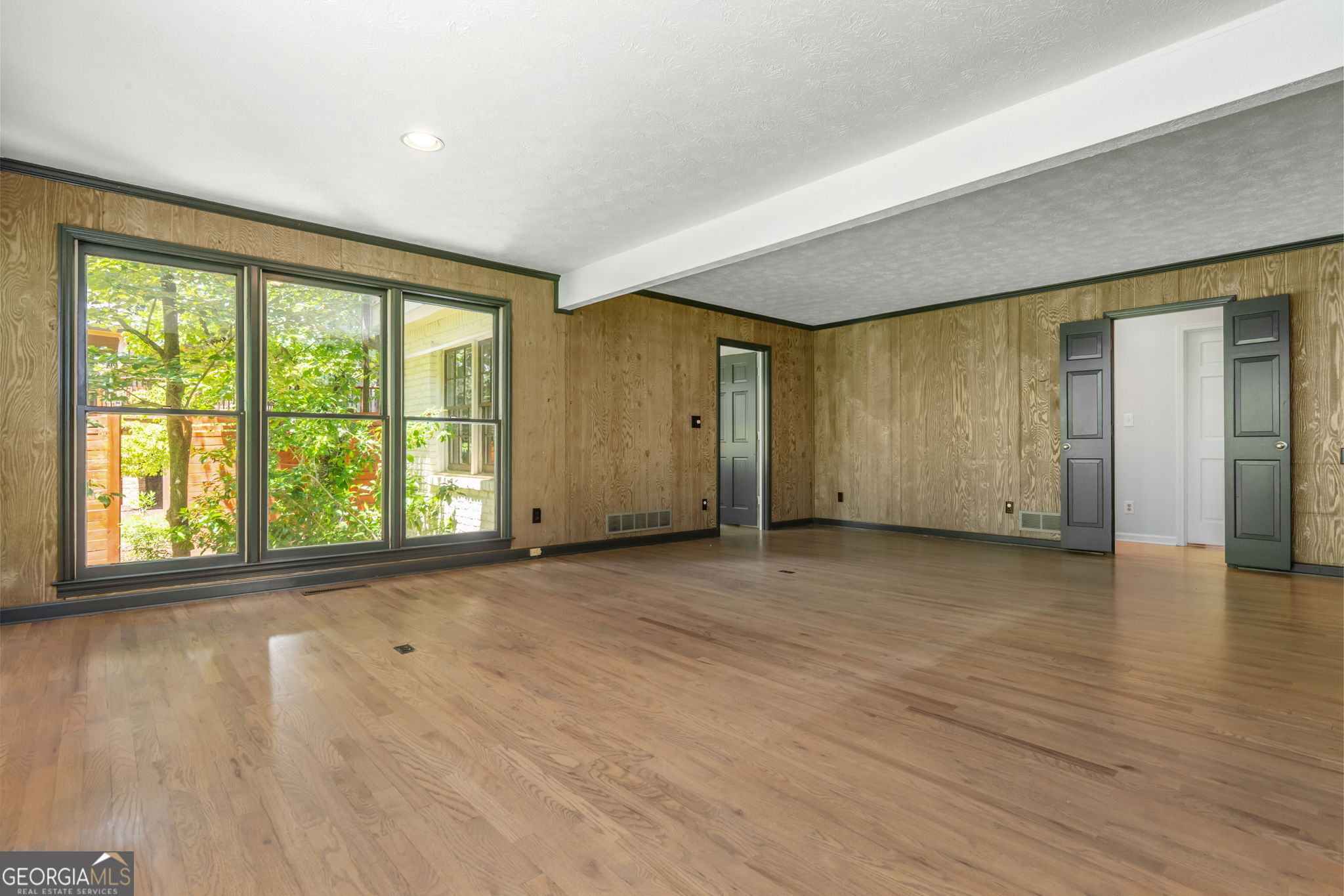 Empty room with hardwood floors, wood-paneled walls, and large windows.
