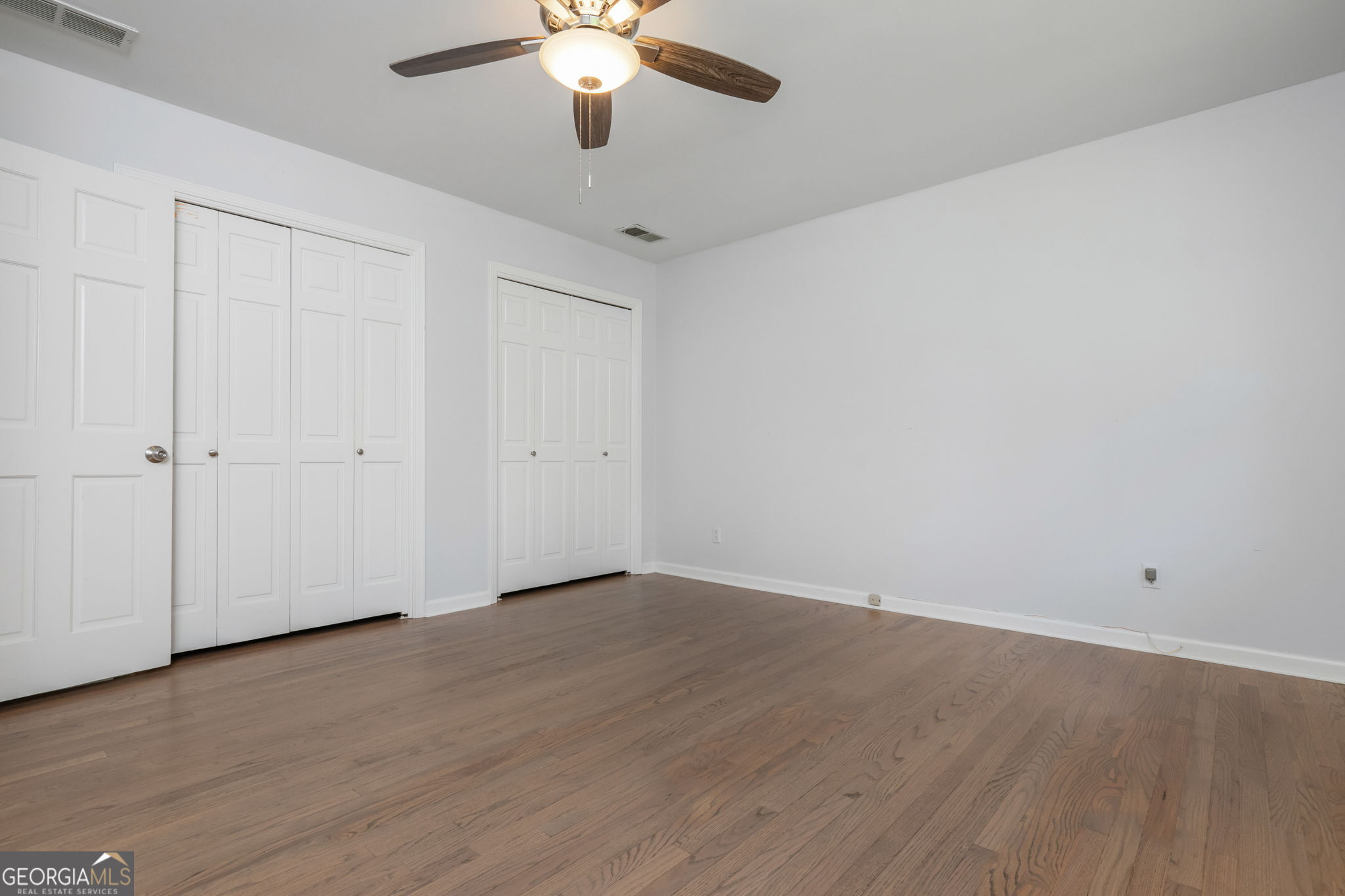 Empty bedroom with hardwood floors, white paneled closet doors, and a ceiling fan with light.