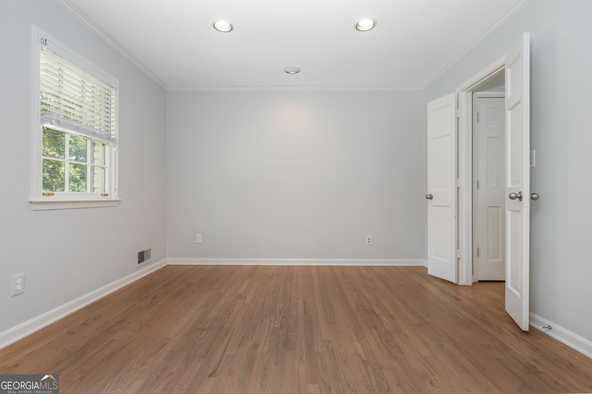 Empty room with hardwood floors, a window with white blinds, and open doors.