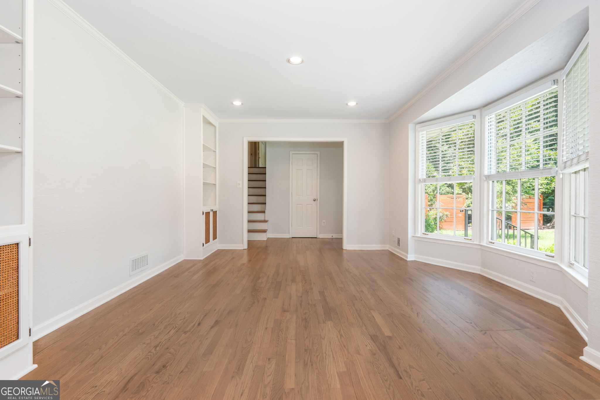 Vacant living room with hardwood floors, built-in shelving, and large windows overlooking greenery.
