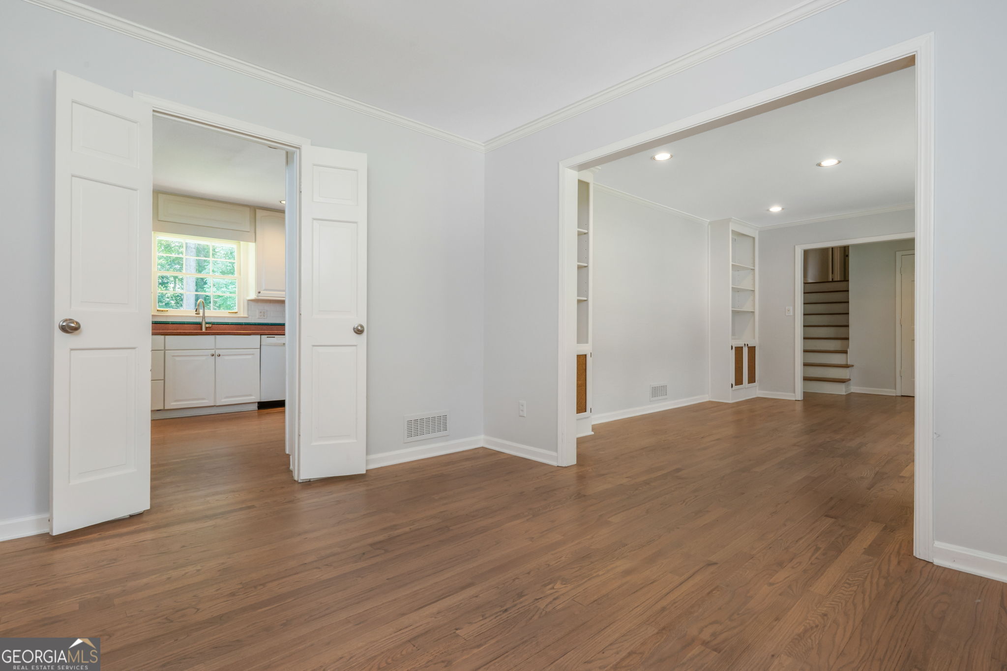 Empty room with hardwood floors, open kitchen doorway, built-in shelves, and stairs.