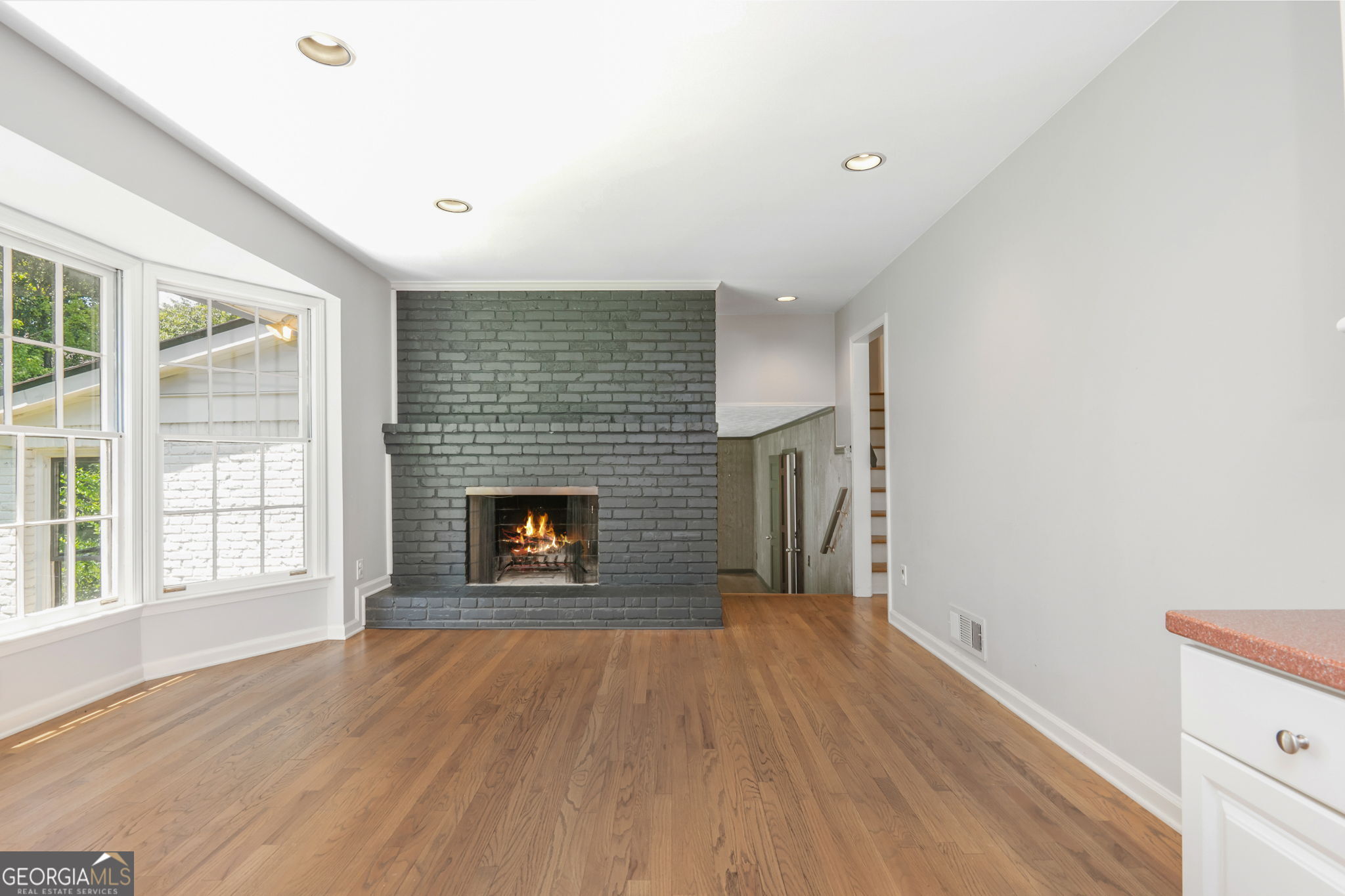 Vacant living room with hardwood floors, tall windows, and a dark brick fireplace with a fire.