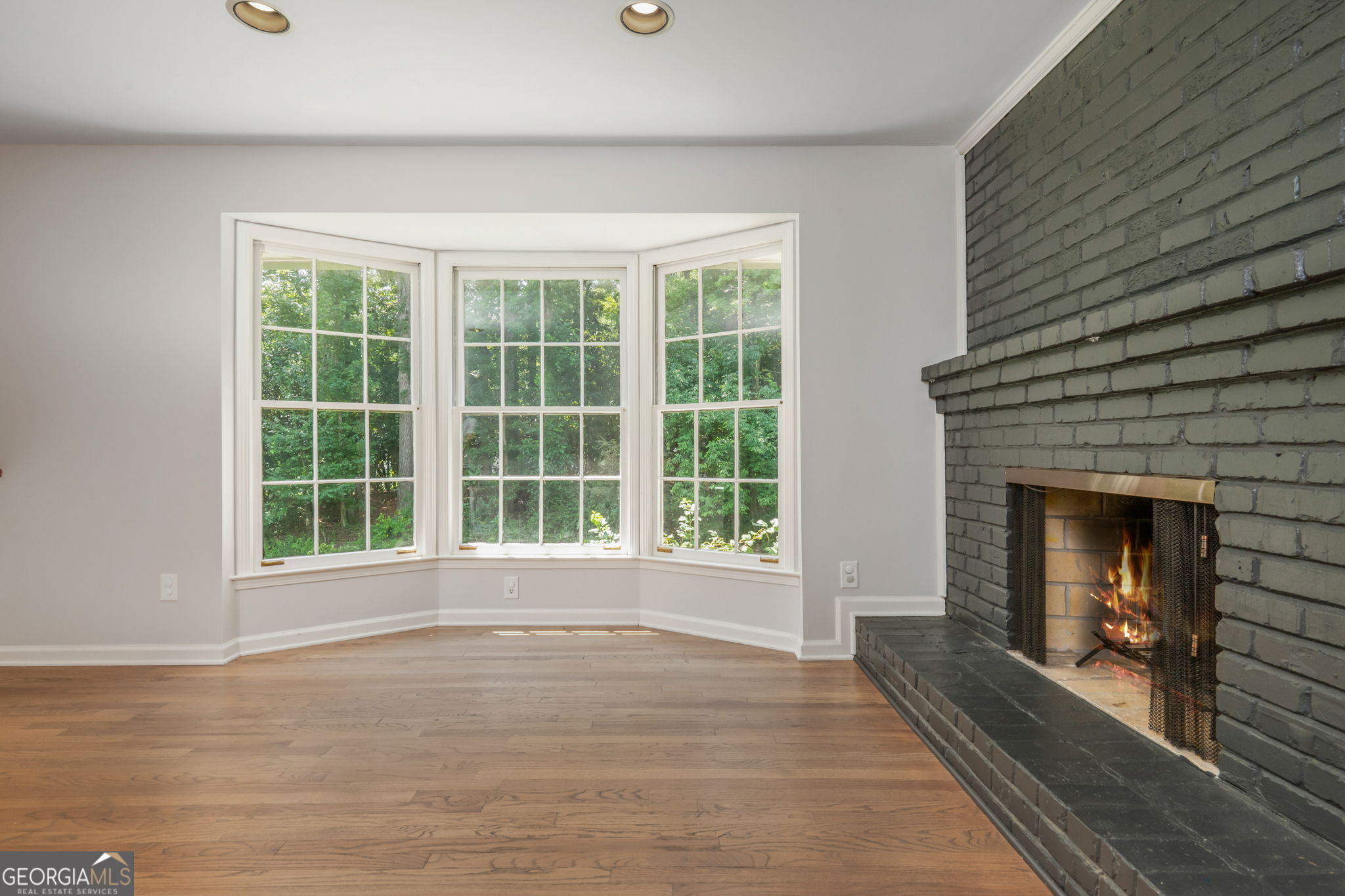 Empty living room with a bay window, hardwood floors, and a lit dark brick fireplace.