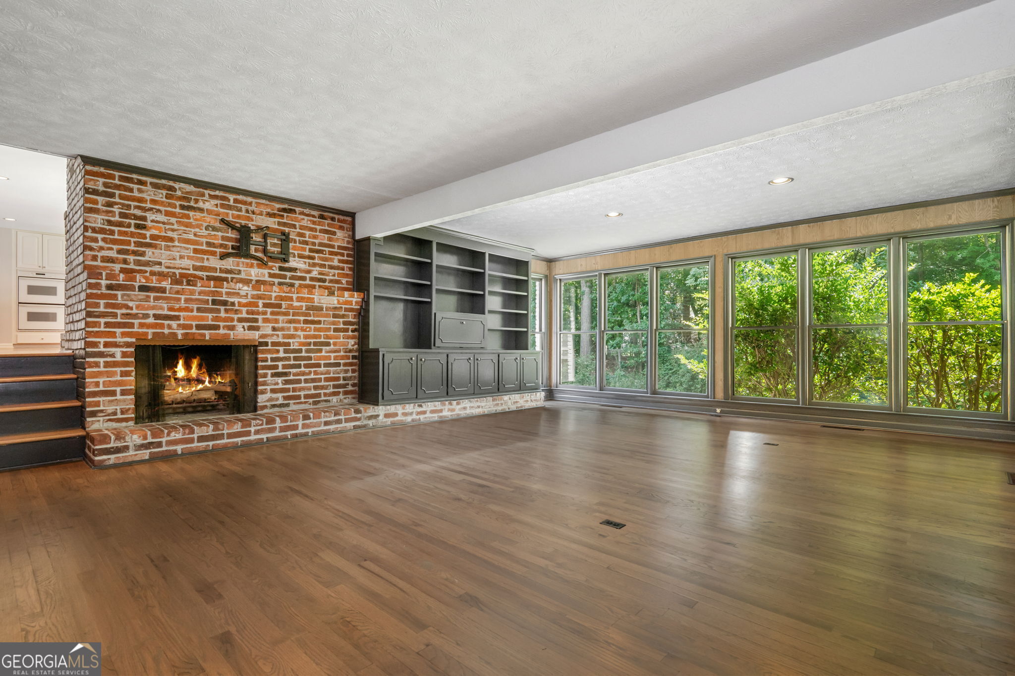 Vacant living room with a brick fireplace, built-in shelving, hardwood floors, and large windows overlooking trees.