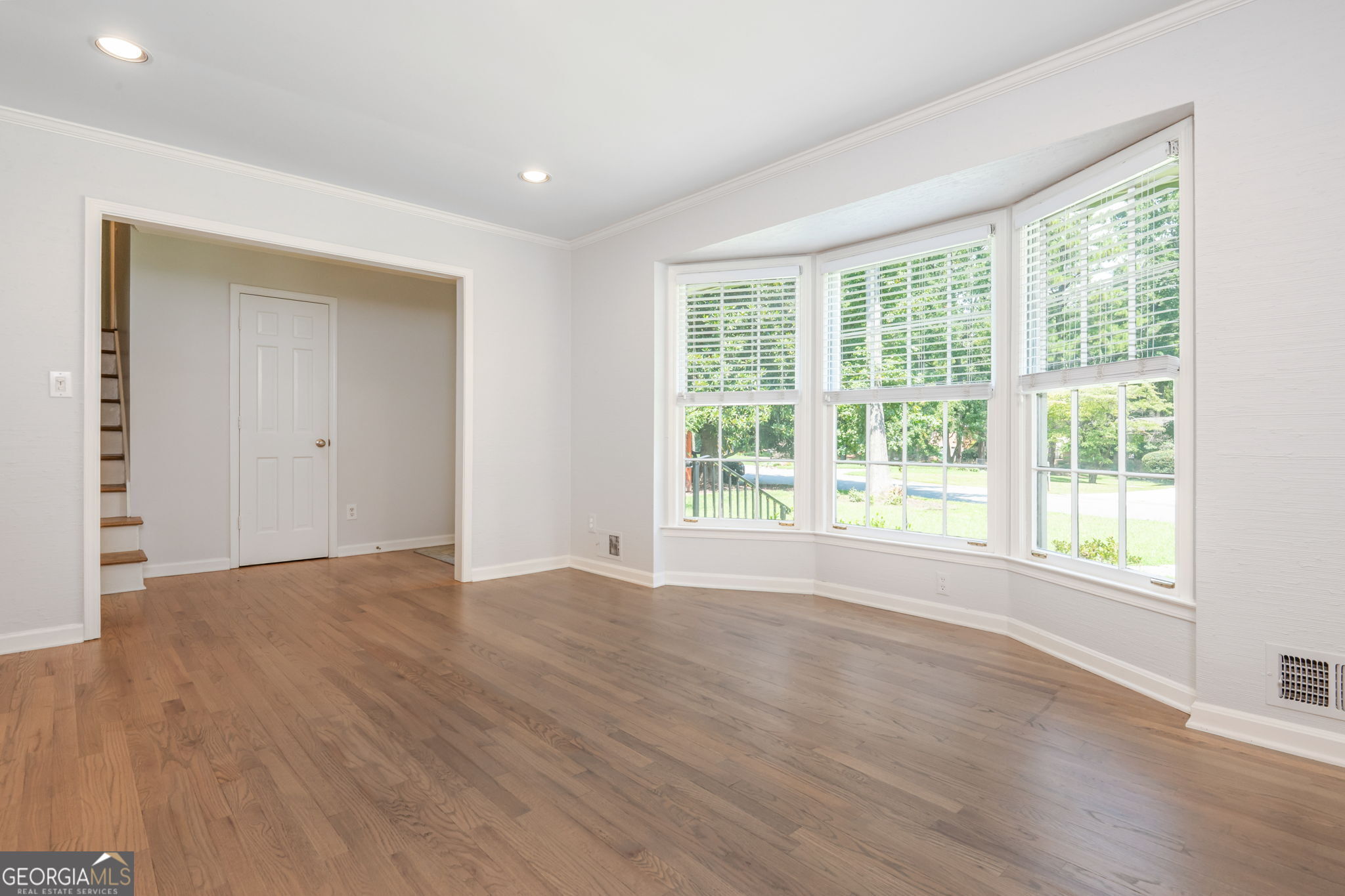 Empty living room with hardwood floors and a large bay window overlooking greenery.