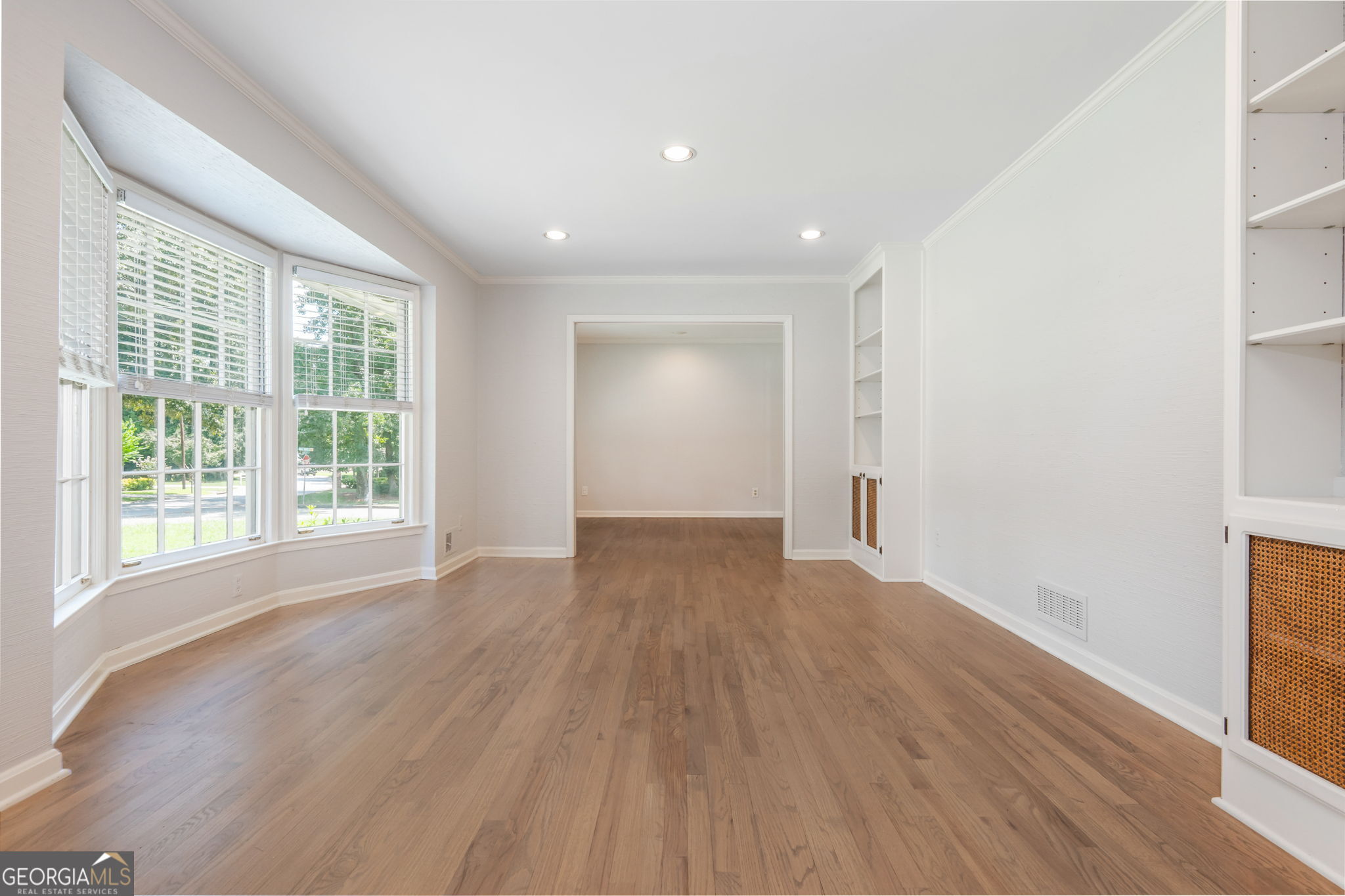 Empty living room with hardwood floors, large windows, and built-in shelving.