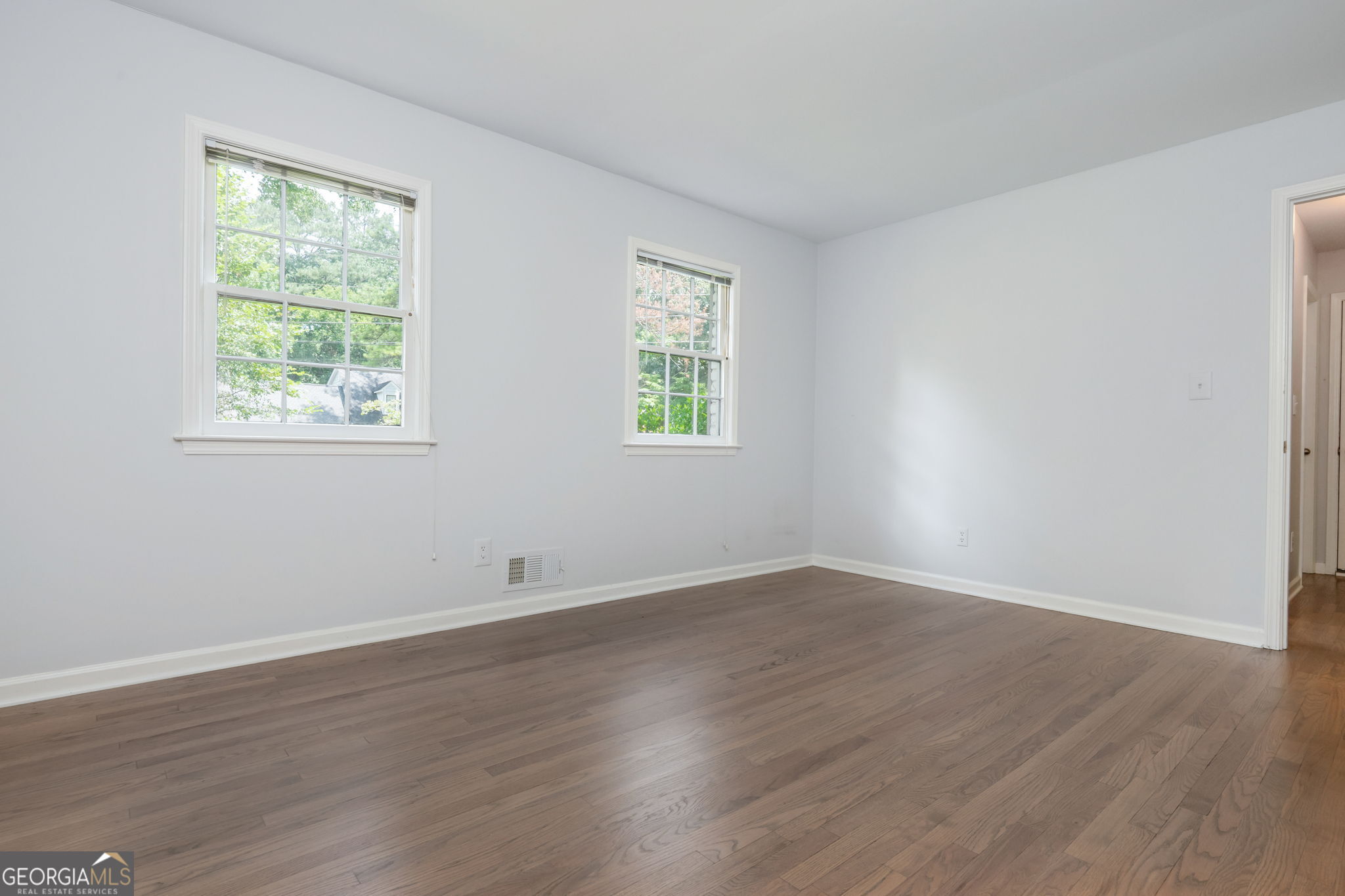 Empty room with hardwood floors, pale walls, and two windows overlooking greenery.
