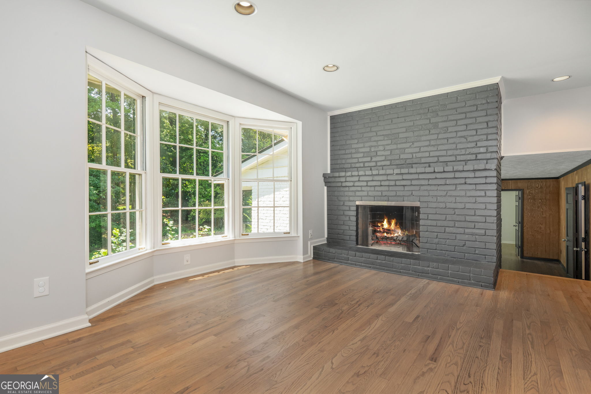 Empty living room with hardwood floors, a gray brick fireplace, and three large windows.