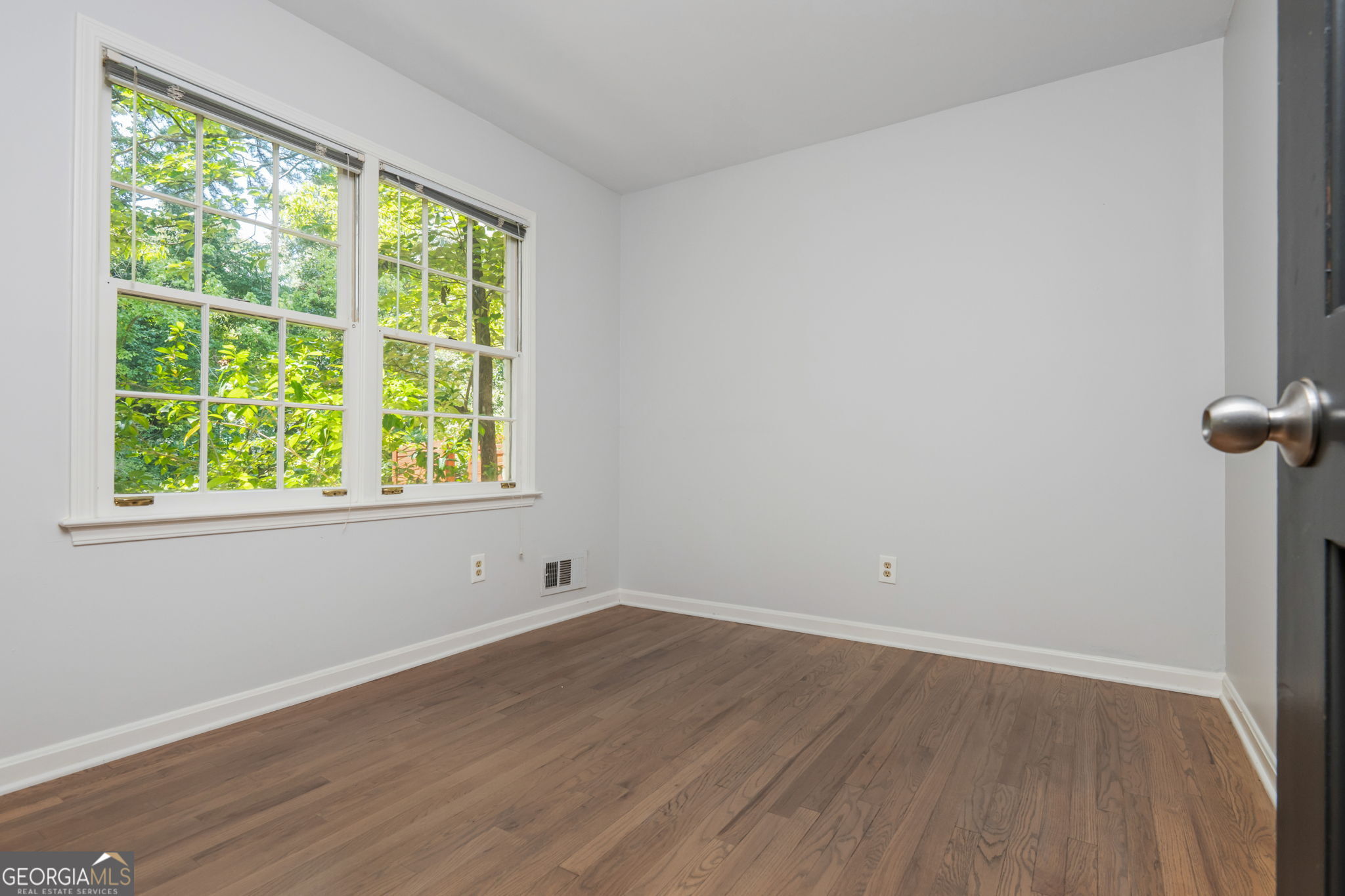 Empty room with hardwood floors and two large windows overlooking leafy greenery.