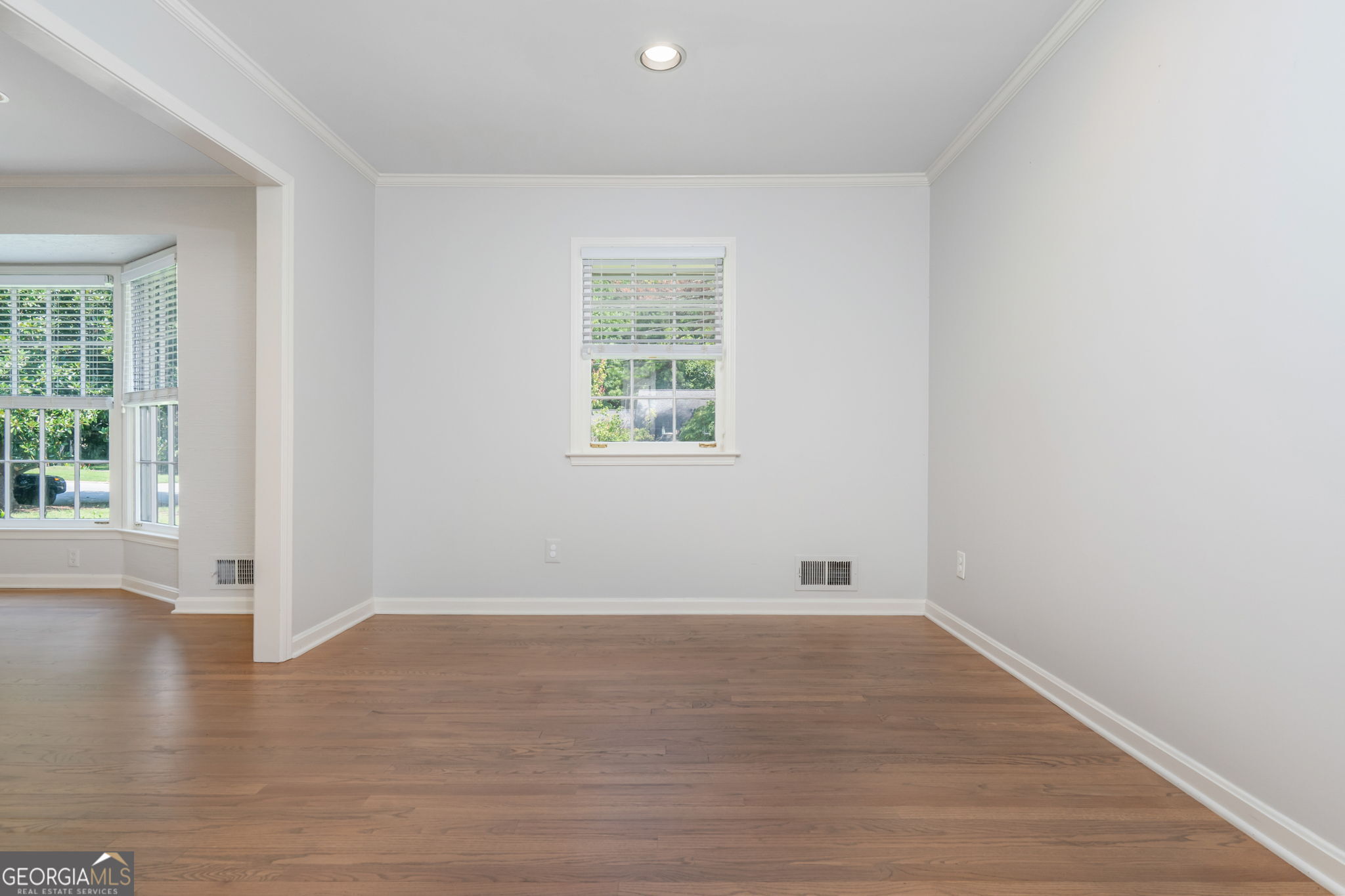 Vacant room with hardwood floors and a window, opening to a bright bay-window area.