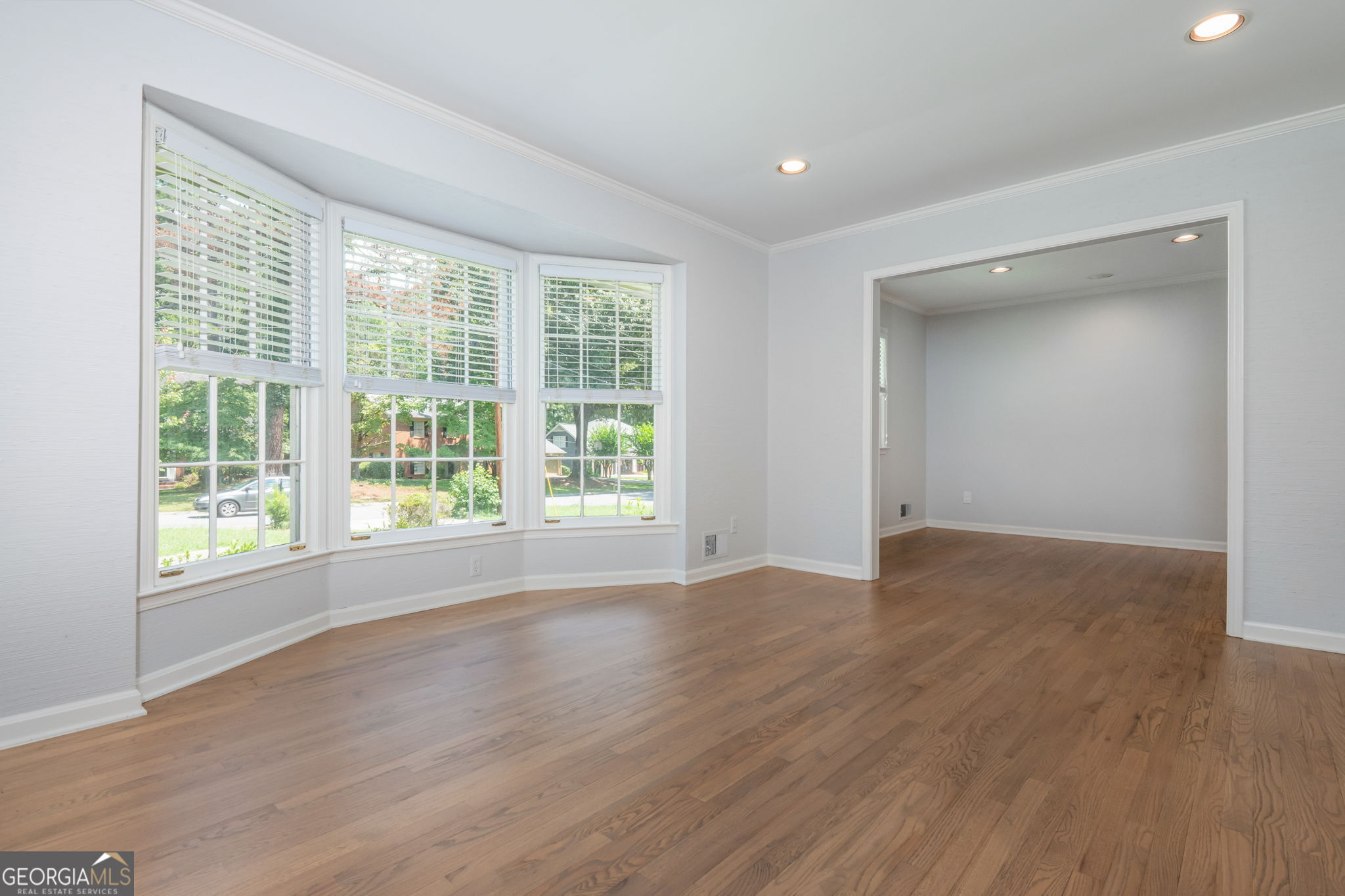 Empty living room with hardwood floors, three wide windows with blinds, and a large opening to an adjoining room.