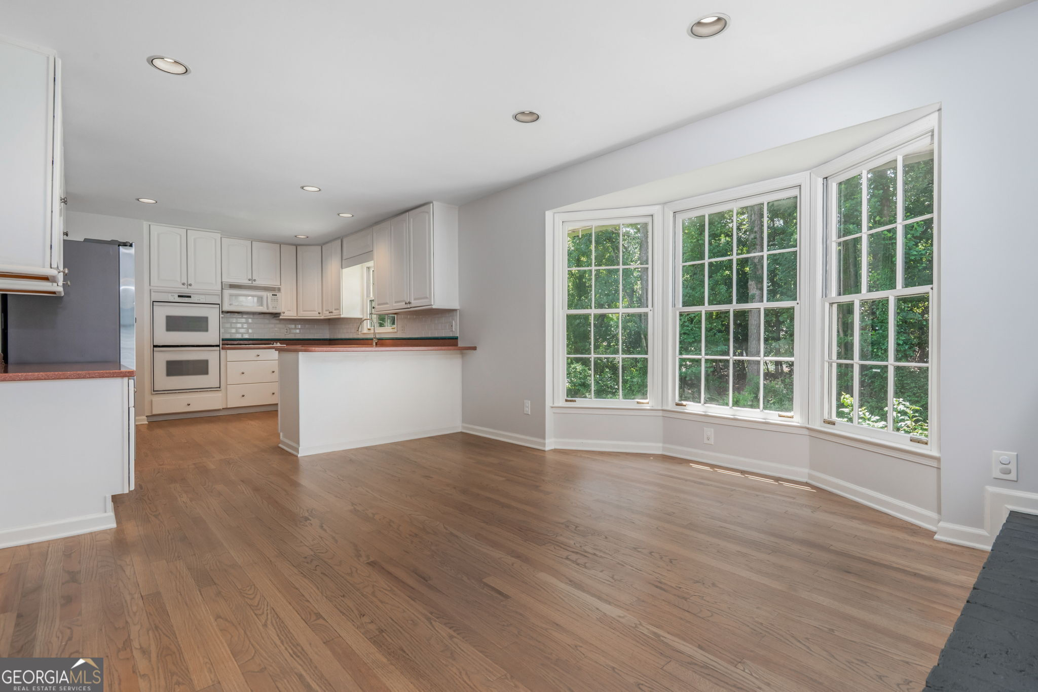 Open kitchen and dining area with hardwood floors, white cabinets, and a bay window overlooking trees.
