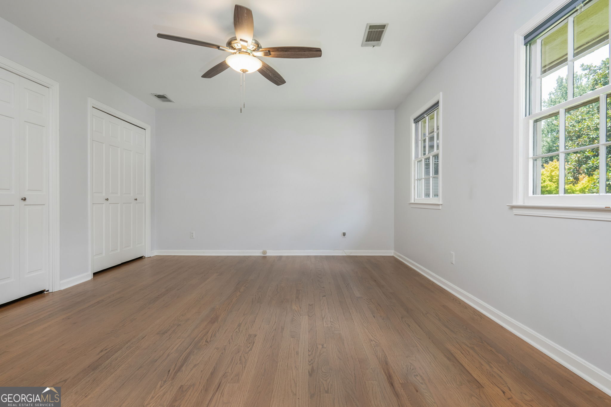 Empty bedroom with hardwood floors, double closets, a ceiling fan, and two windows.