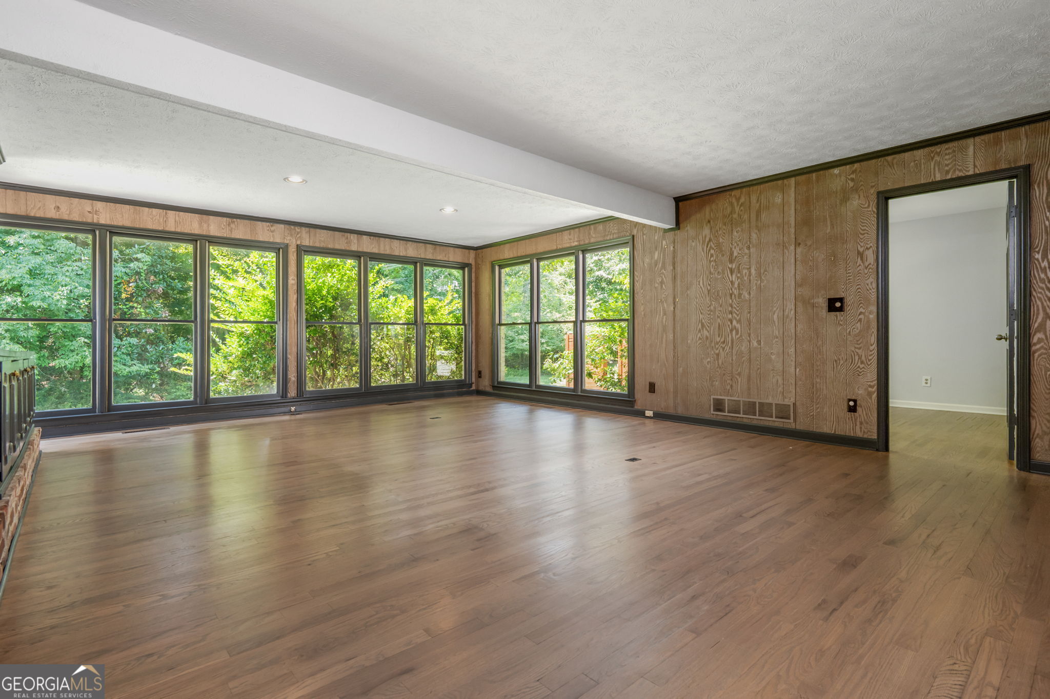 Empty room with hardwood floors, wood-paneled walls, and large windows overlooking greenery.
