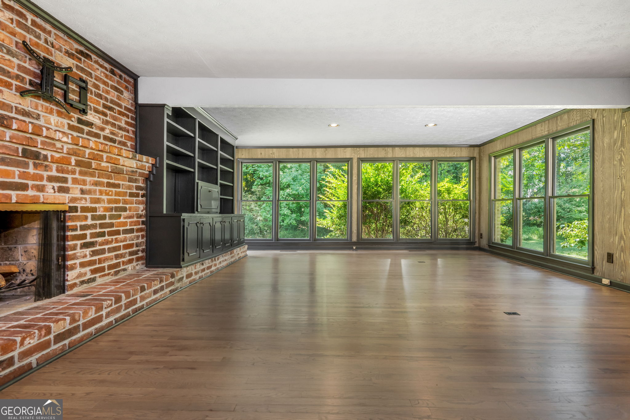 Vacant living room with a brick fireplace, built-in shelving, and broad windows overlooking greenery.