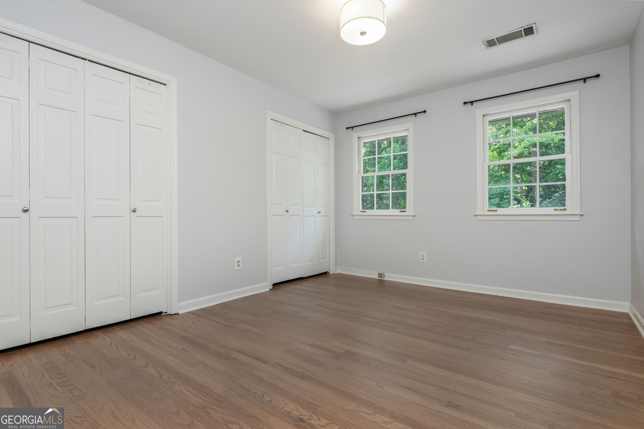 Empty bedroom with hardwood floors, two white closets, and windows overlooking trees.