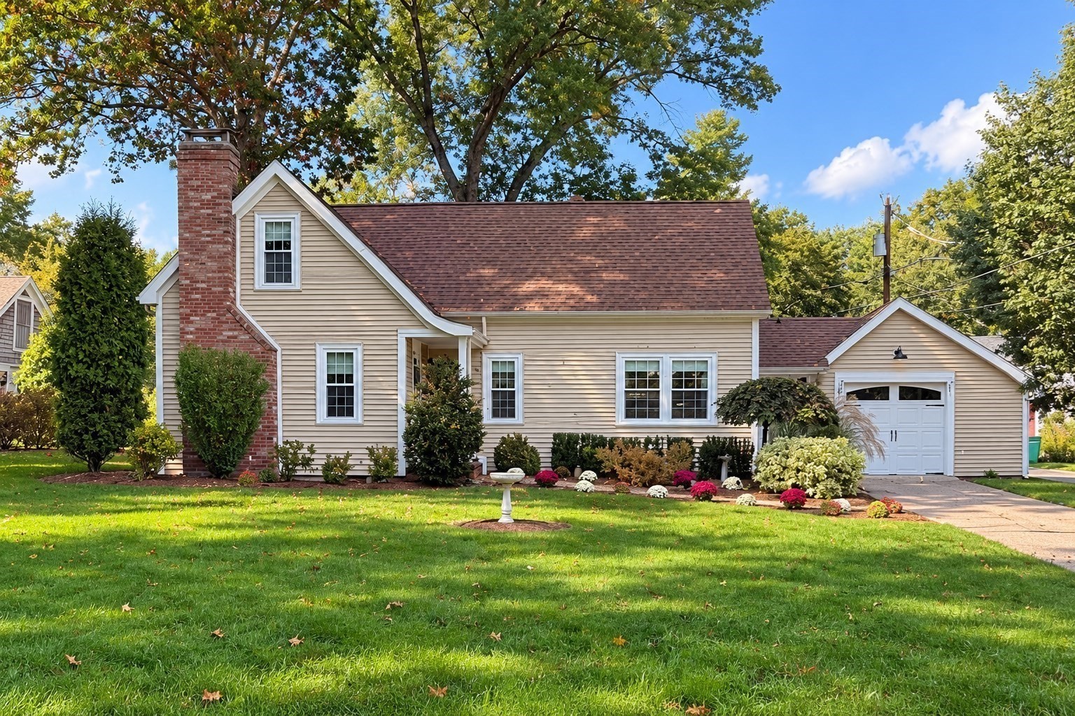 Beige-sided house with a brick chimney, attached garage, and landscaped front lawn.