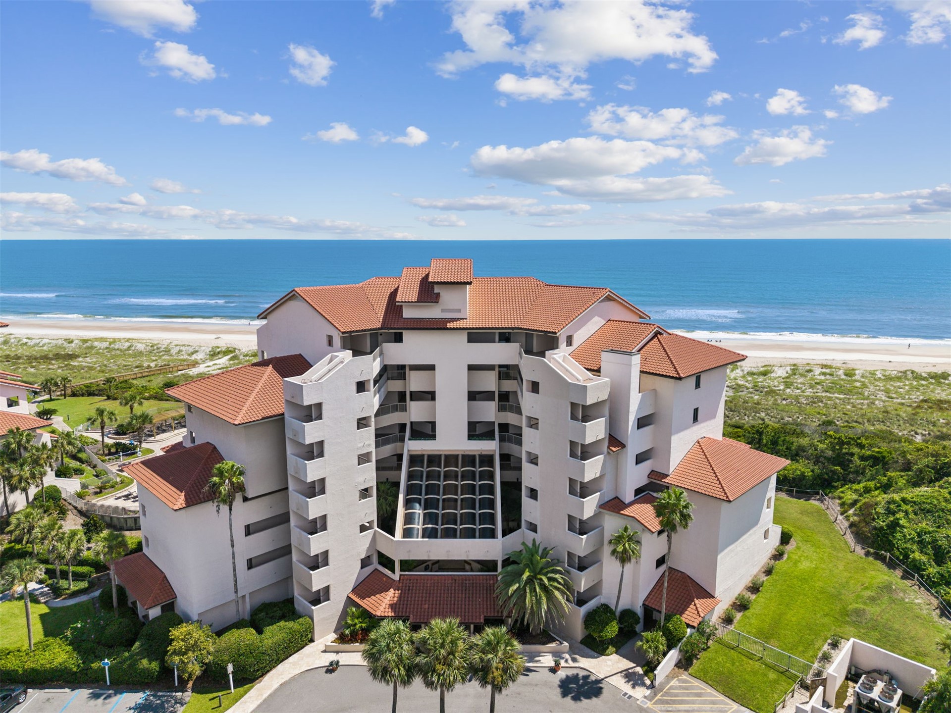 Aerial view of a beachfront condominium with terracotta roofs, palm trees, sandy beach, and ocean.