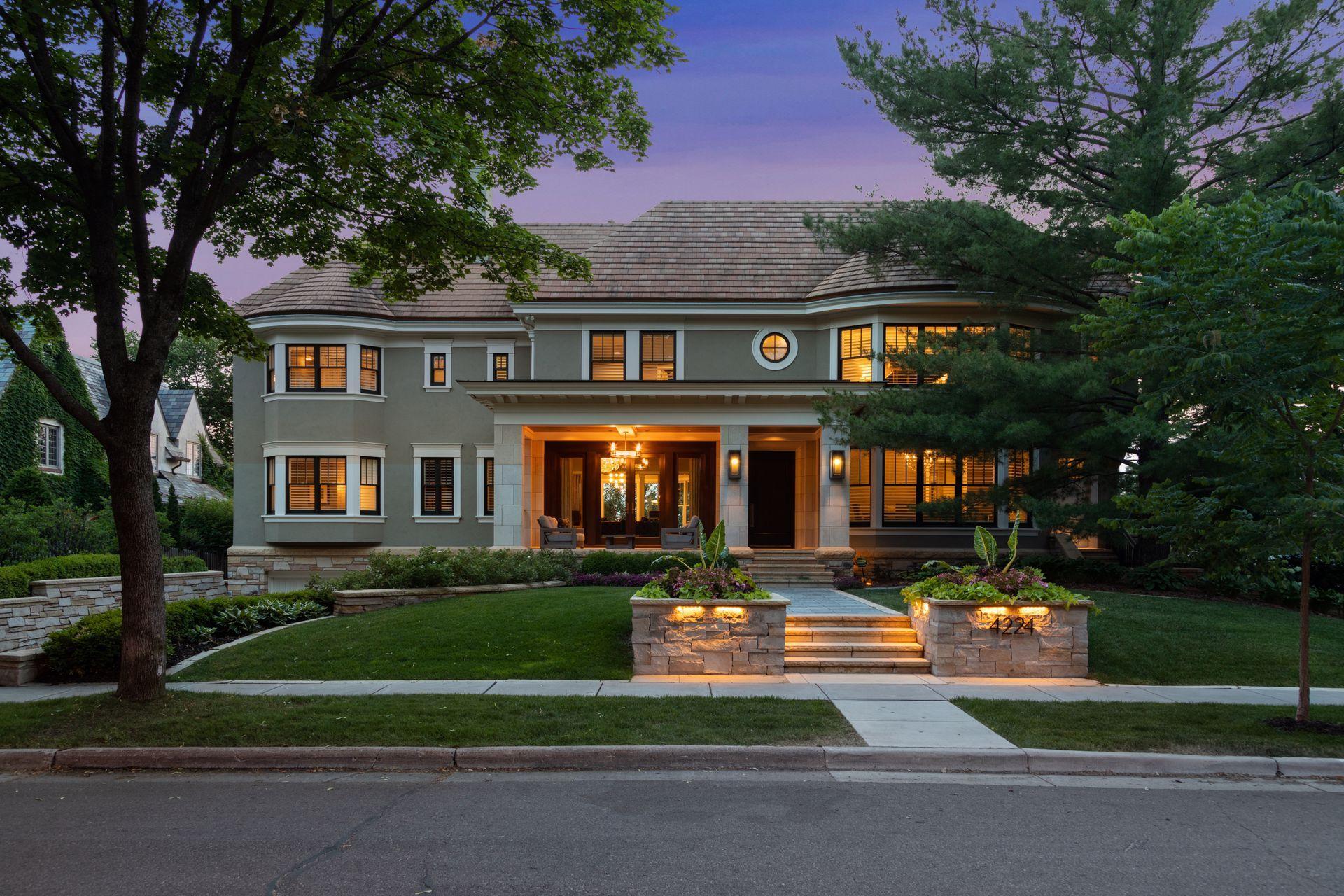 Large gray two-story house with a columned entrance, warmly lit windows, and landscaped front steps.
