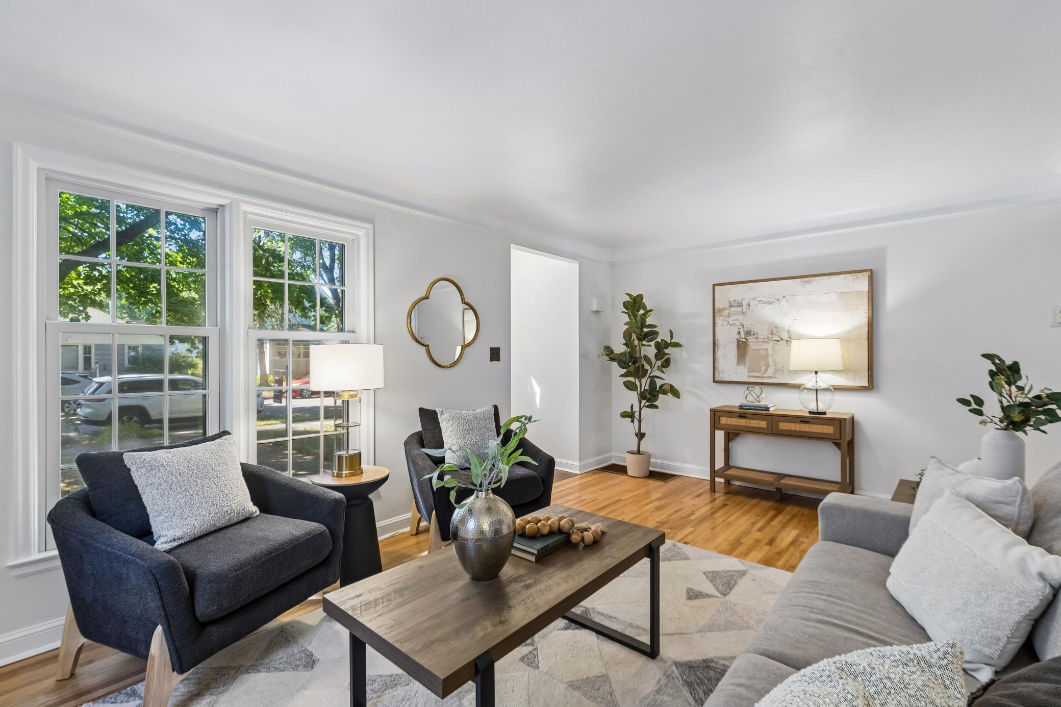 Staged living room with large leafy-window views, hardwood floors, and charcoal and gray seating.