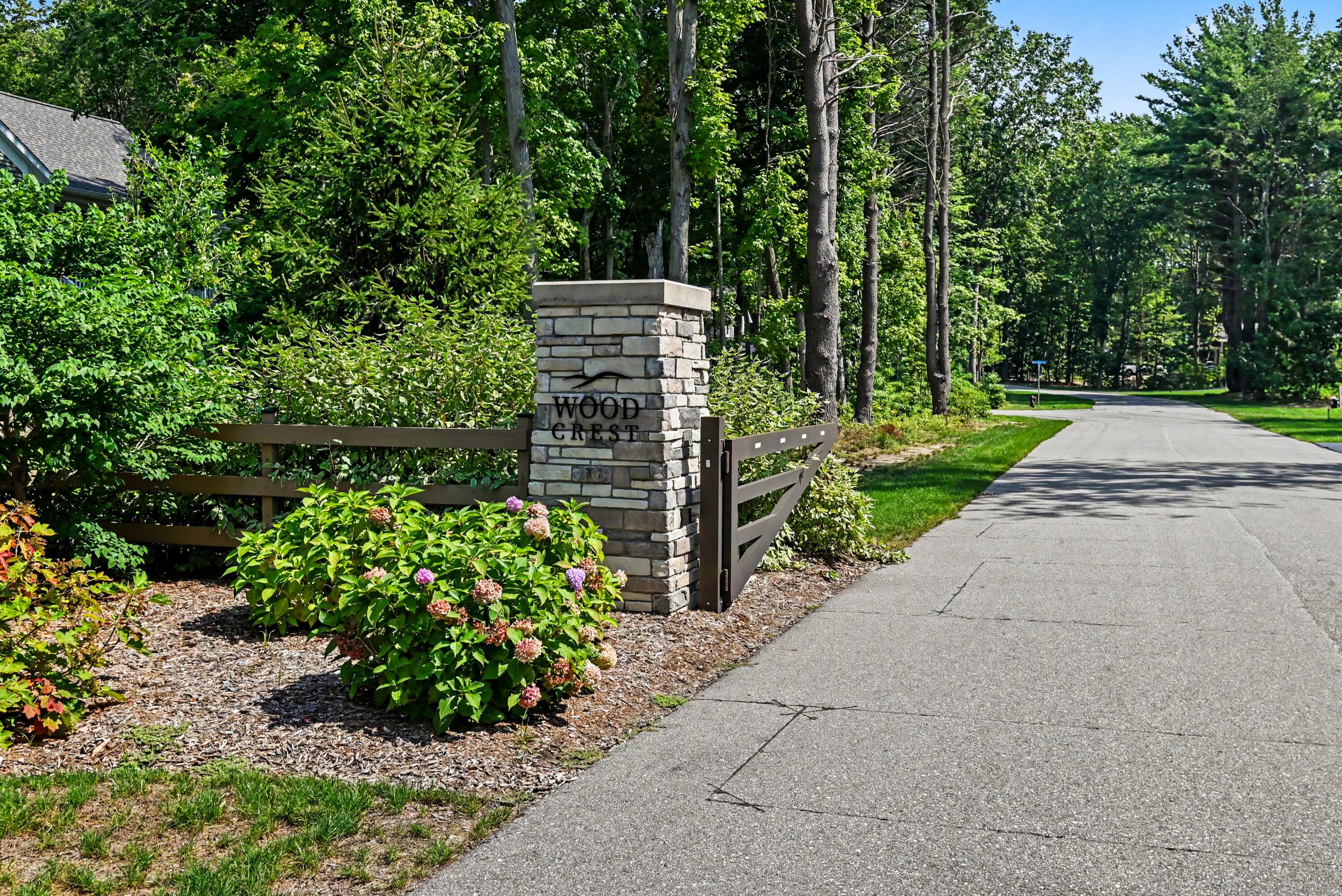 Wood Crest entrance sign beside flowering shrubs and a tree-lined road.