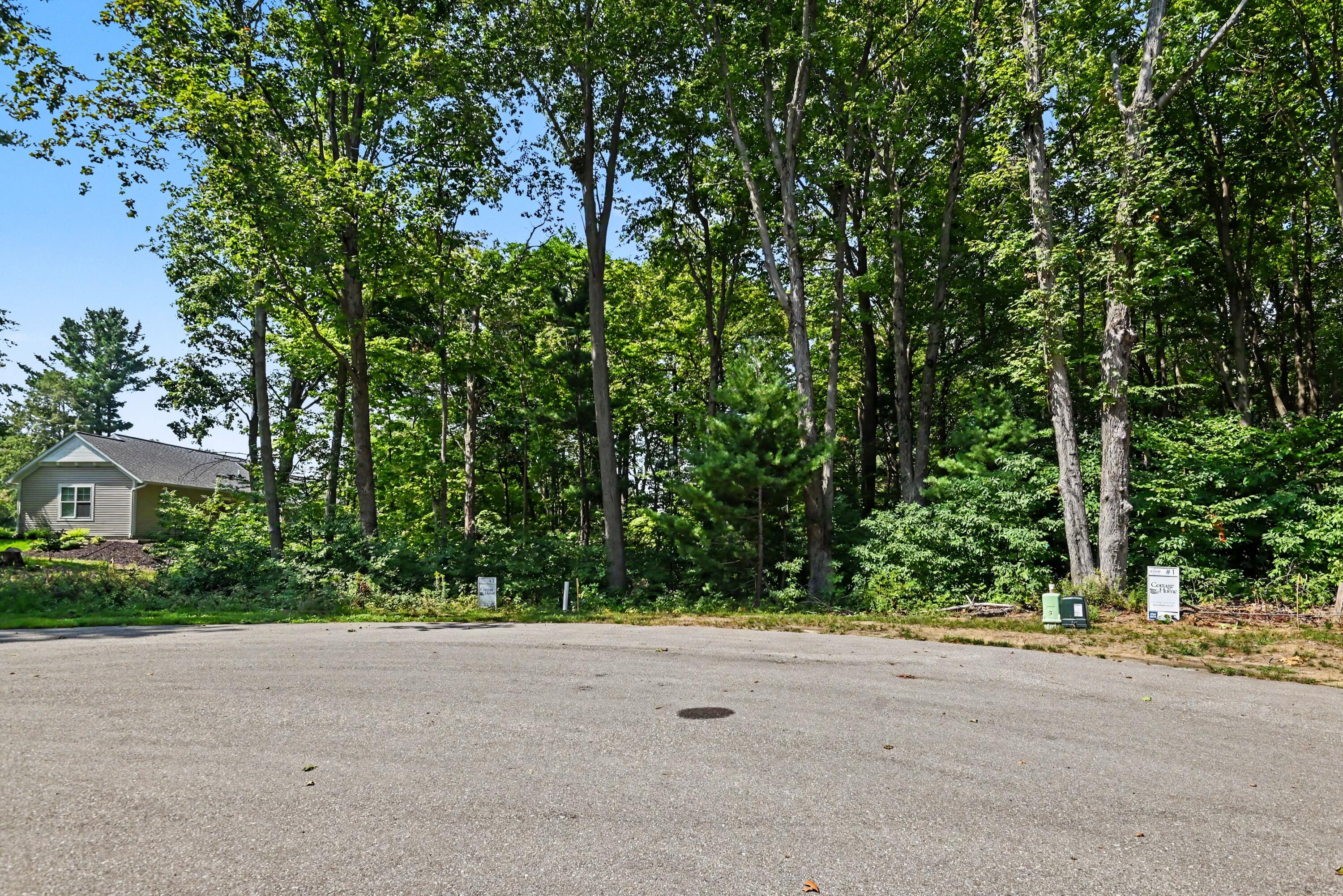 Wooded property lot along a paved road, marked by Cottage Home signs, with a neighboring house at left.