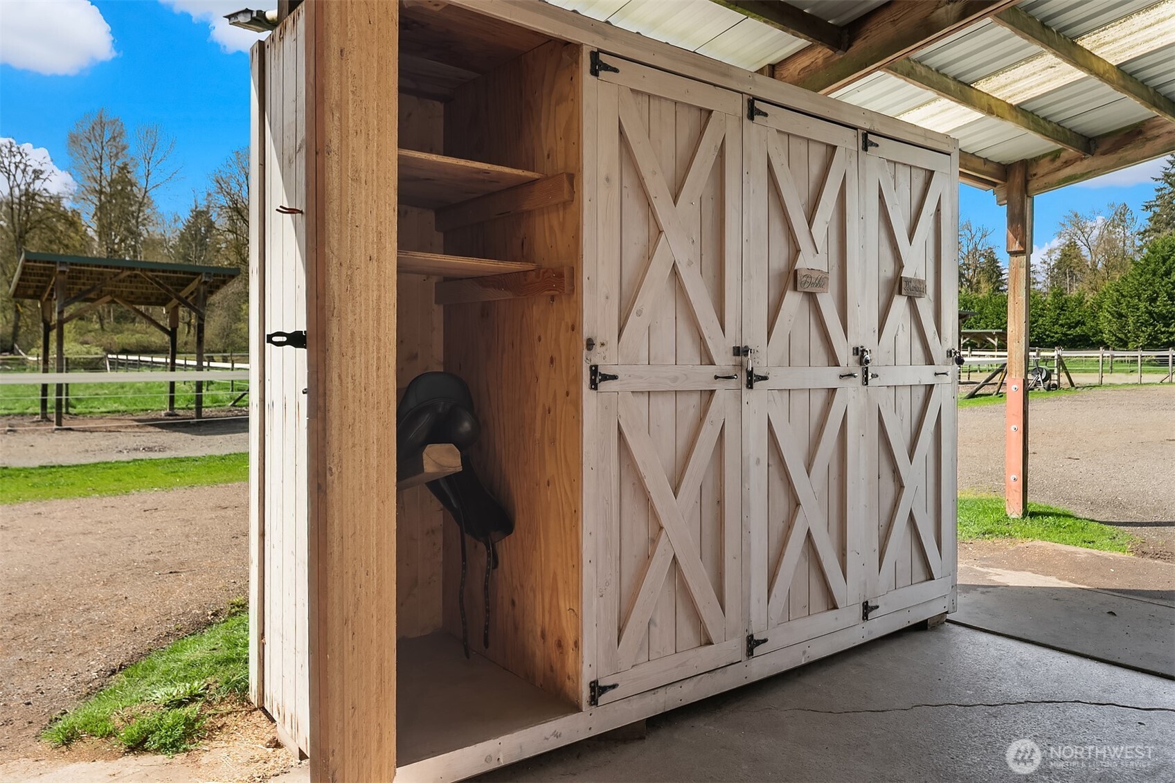 Wooden tack shed with open cross-braced doors and a black saddle inside, beside a fenced paddock.