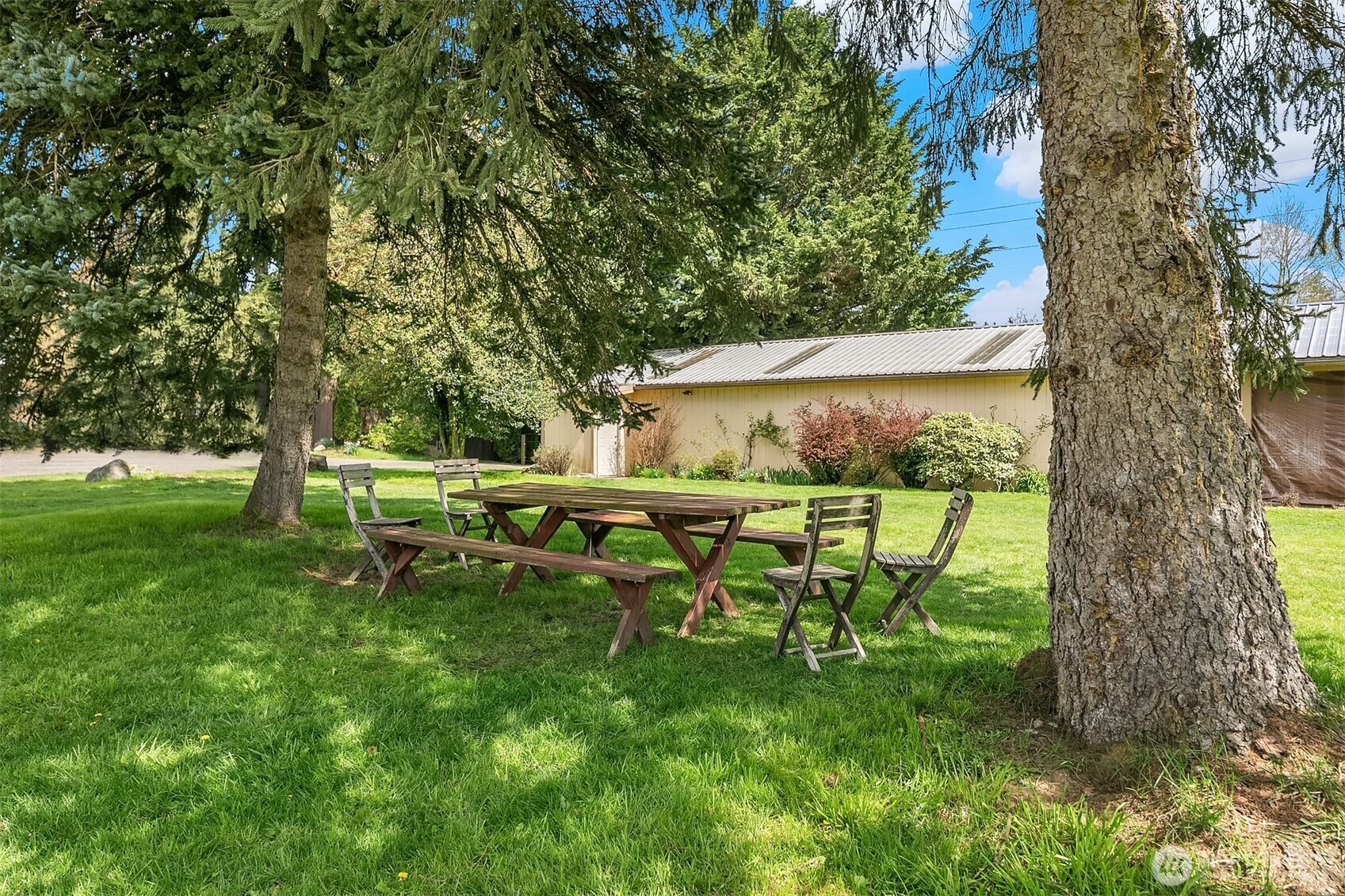 Shaded lawn with a wooden picnic table beside a light-colored outbuilding.