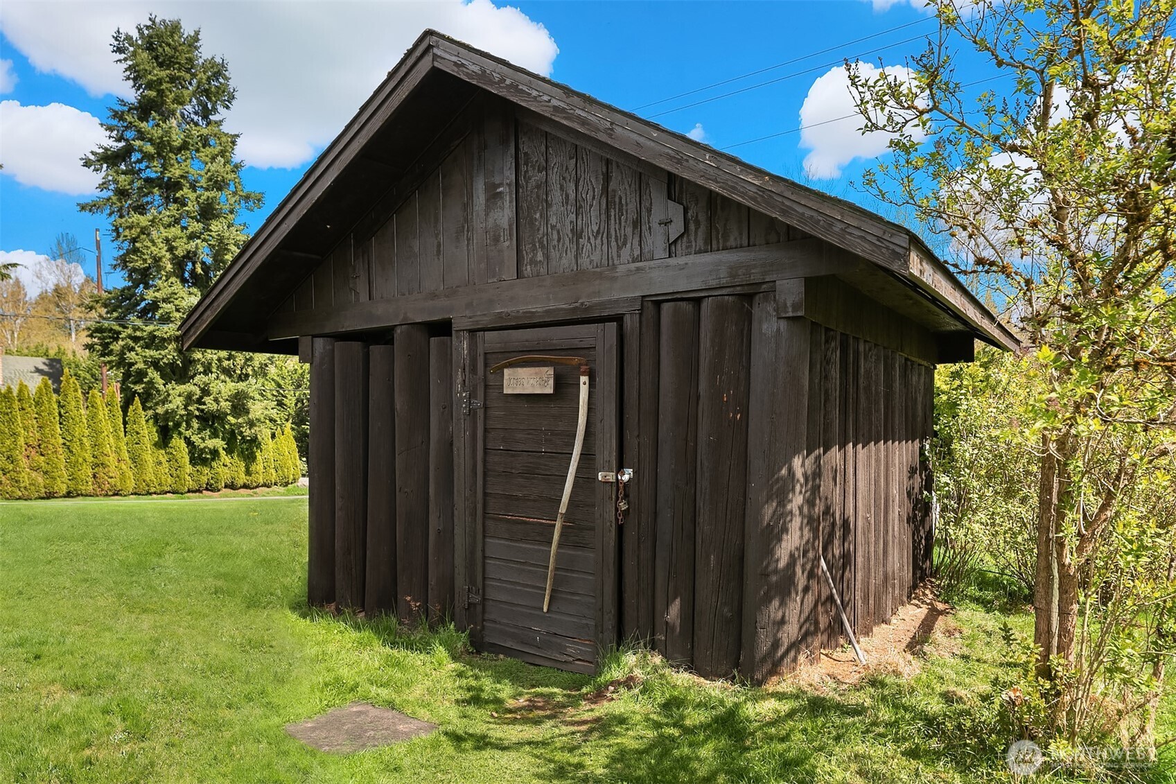 Weathered wooden shed with a steep gabled roof and plank door beside a green lawn.