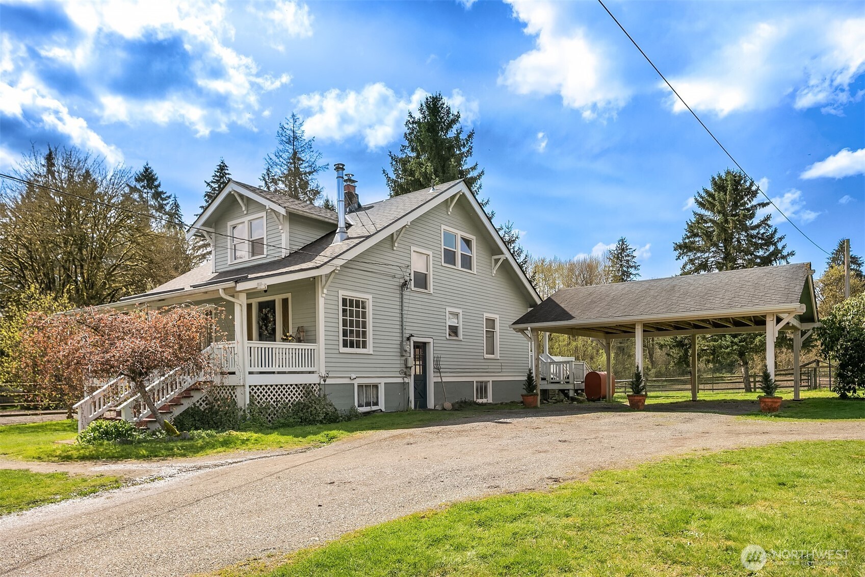 Gray two-story farmhouse with a covered porch and detached carport beside a gravel driveway.