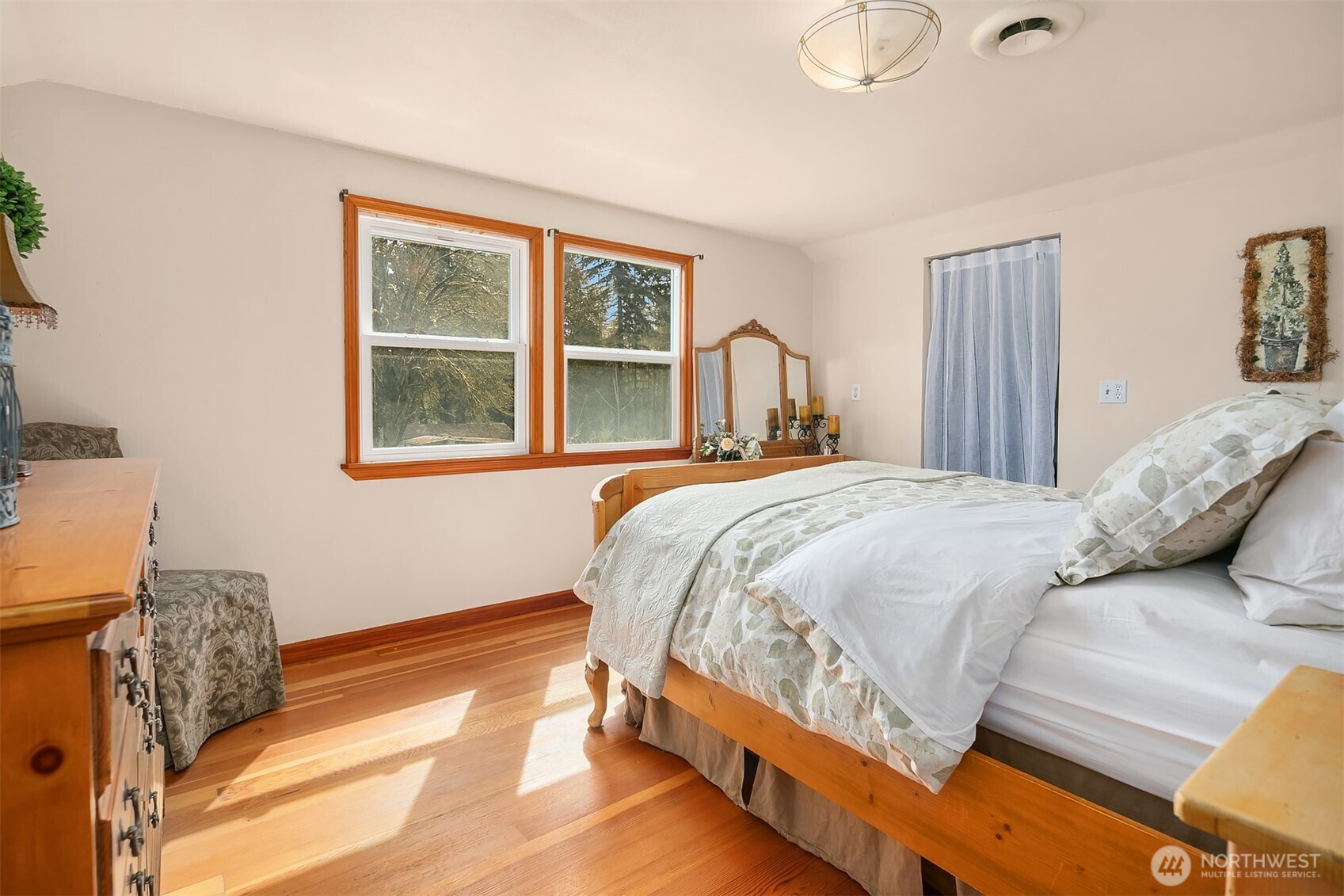 Bedroom with a wooden bed, hardwood floors, and two windows overlooking trees.