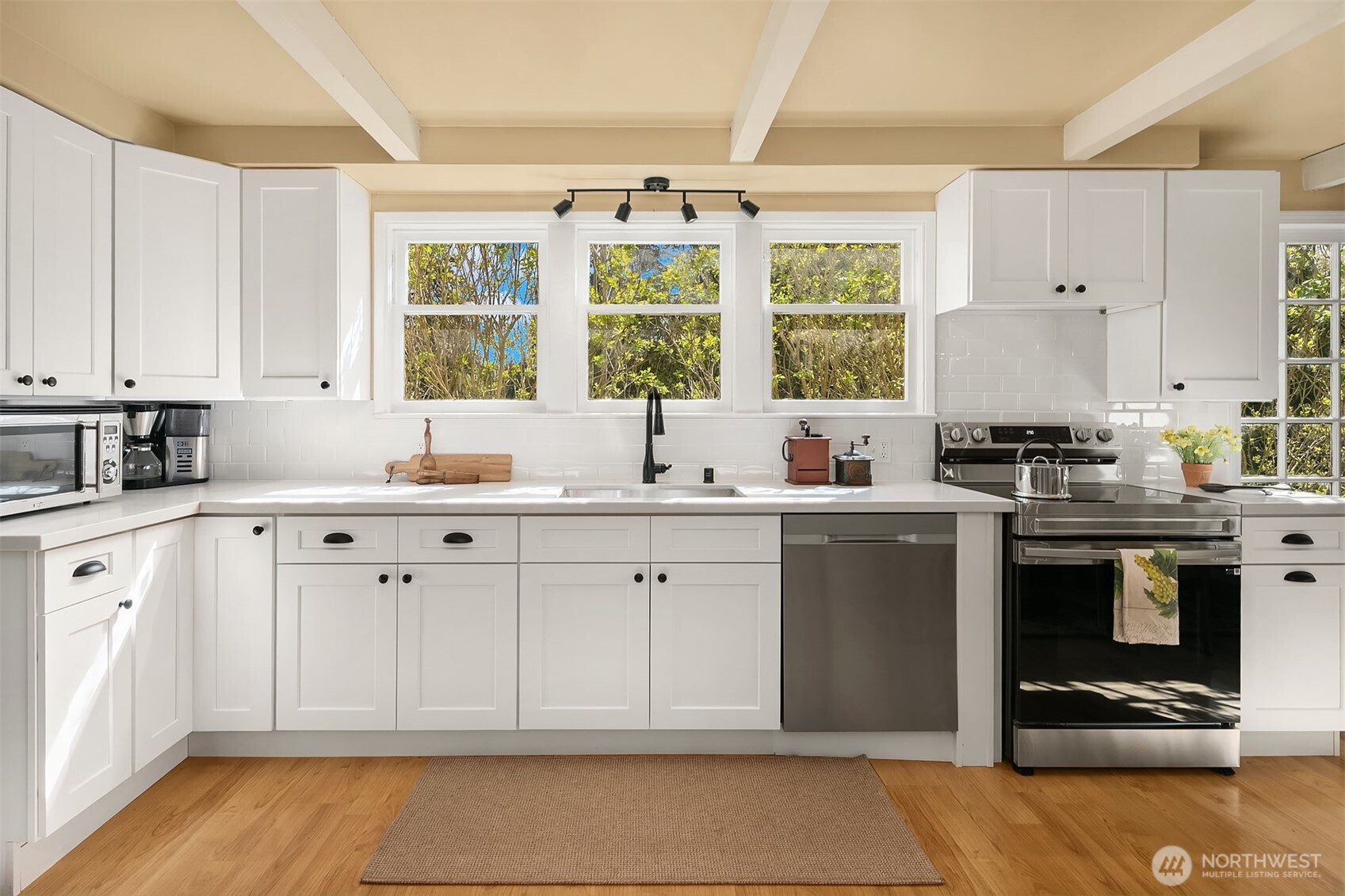 Bright white kitchen with stainless steel appliances, exposed beams, and three windows above the sink.