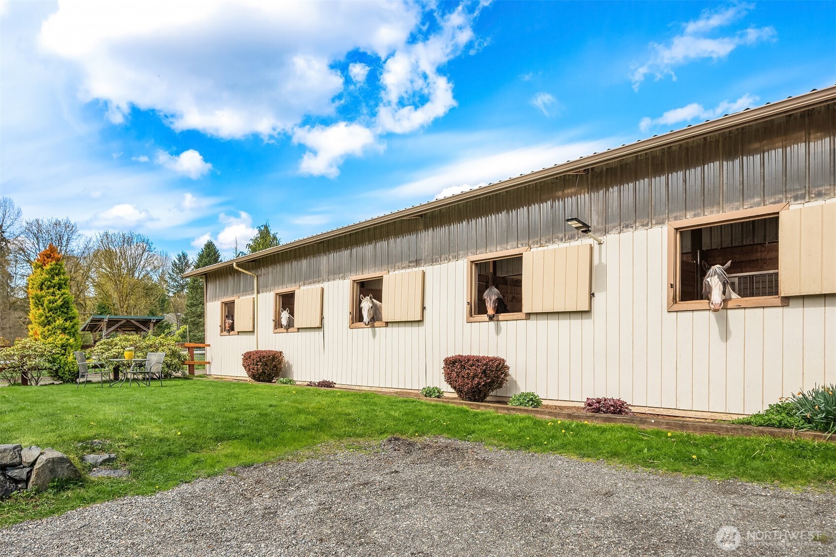 Horse barn with horses looking out of open stall windows beside a gravel drive and lawn.